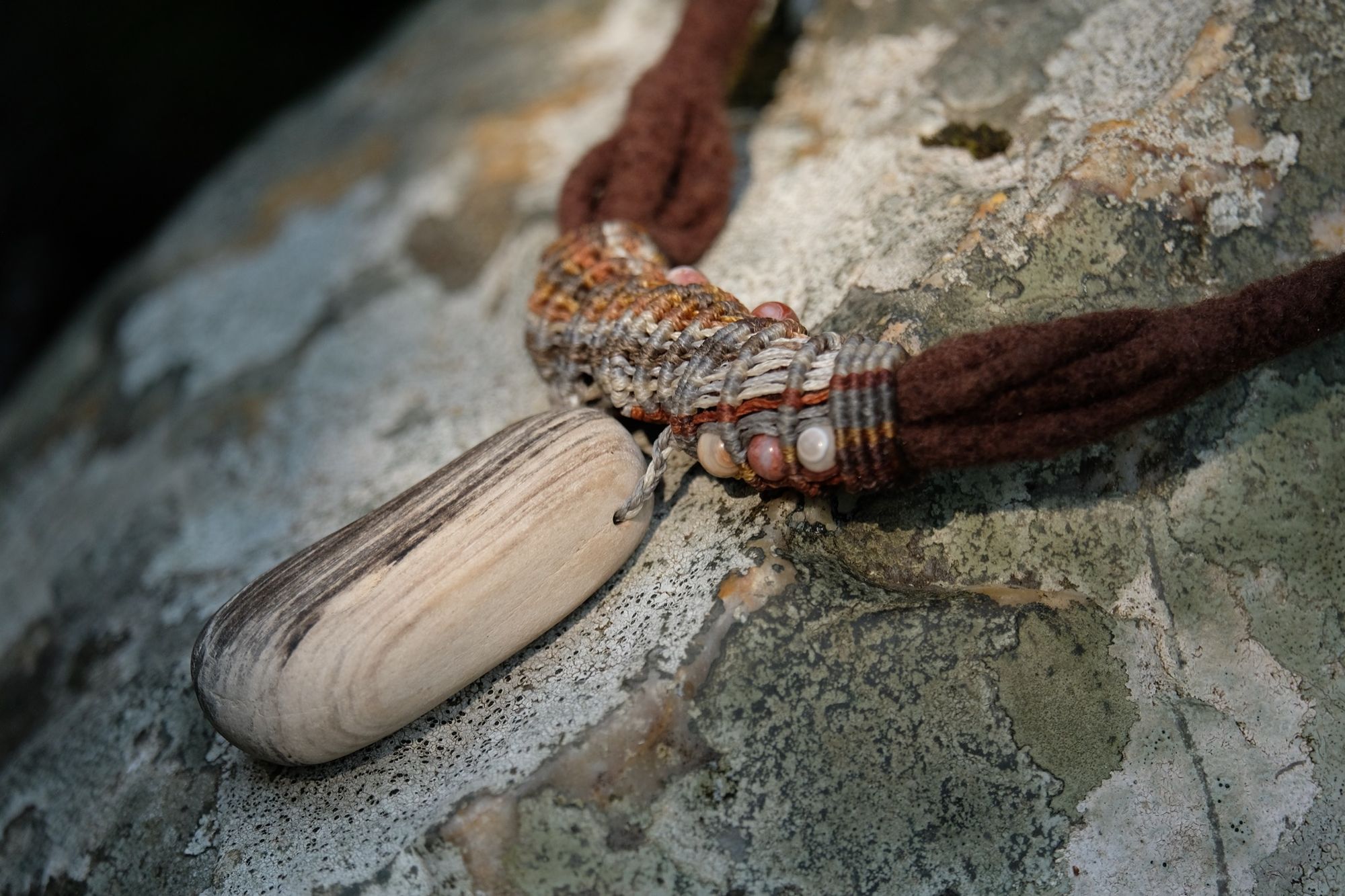 Detail of sculptural necklace made of brown felt, petrified wood and grey-brown knotwork laying on a lichen covered stone