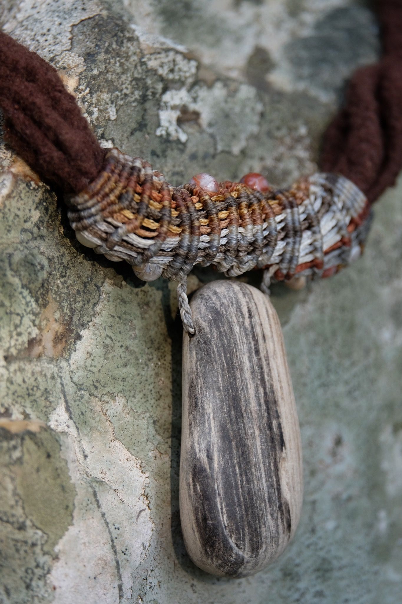 Detail of sculptural necklace made of brown felt, petrified wood and grey-brown knotwork laying on a lichen covered stone