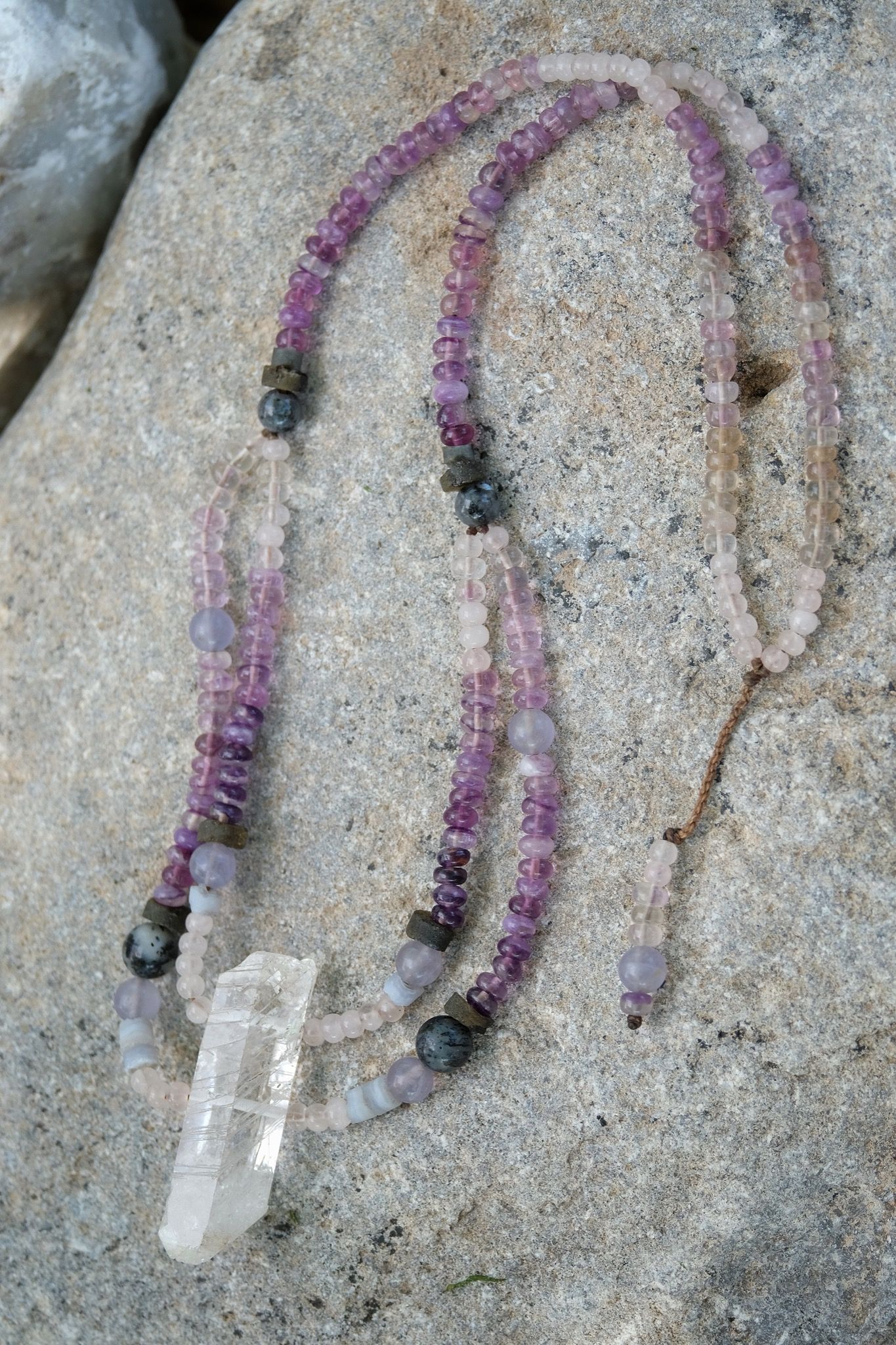 Detail of quartz point, rose quartz, blue chalcedony, purple fluorite and labradorite necklace laying on a rock