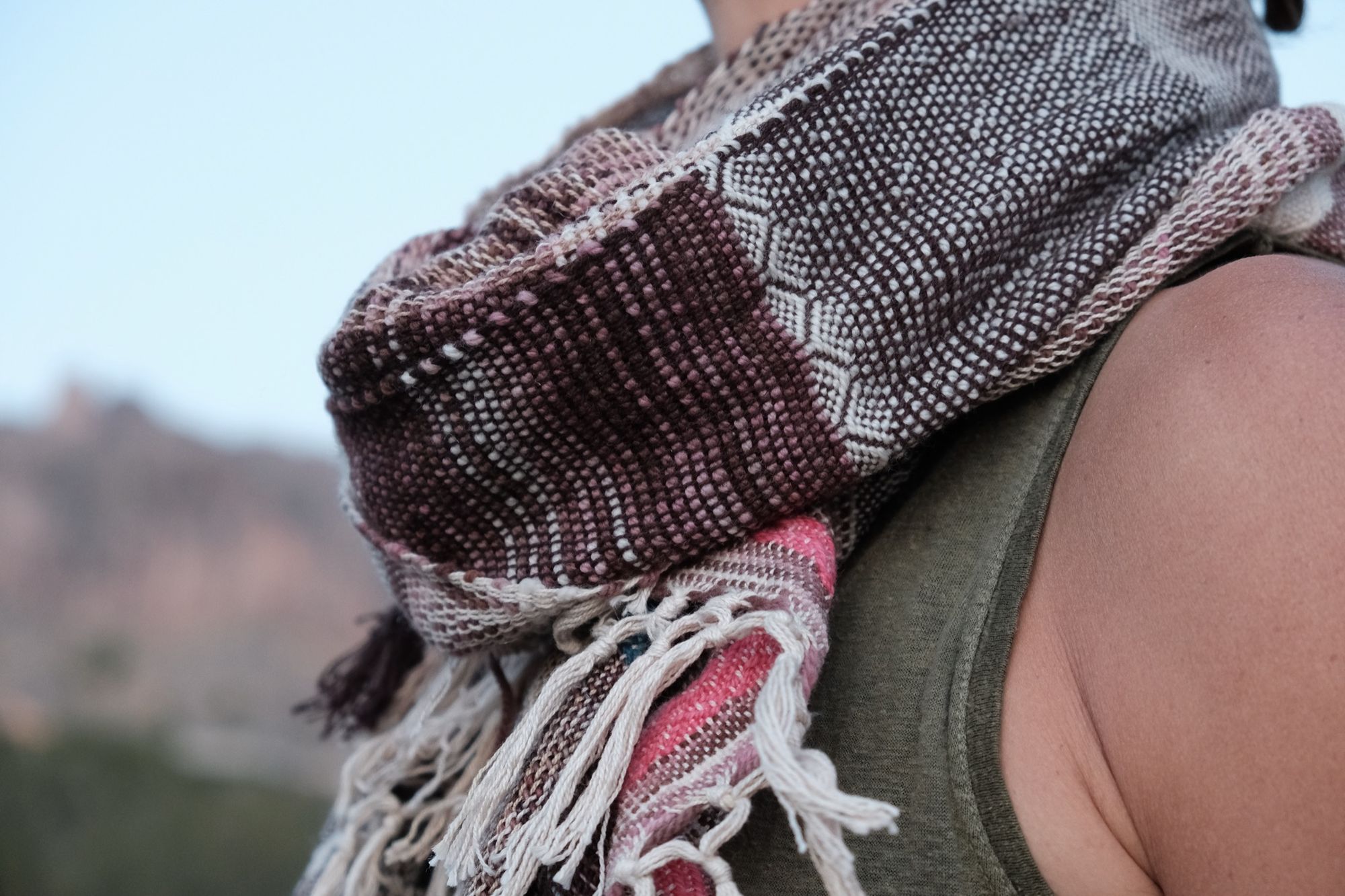 Woman wearing a blue, maroon, white, brown and pink scarf in the desert with cactus, shrubs and mountains in the background. 