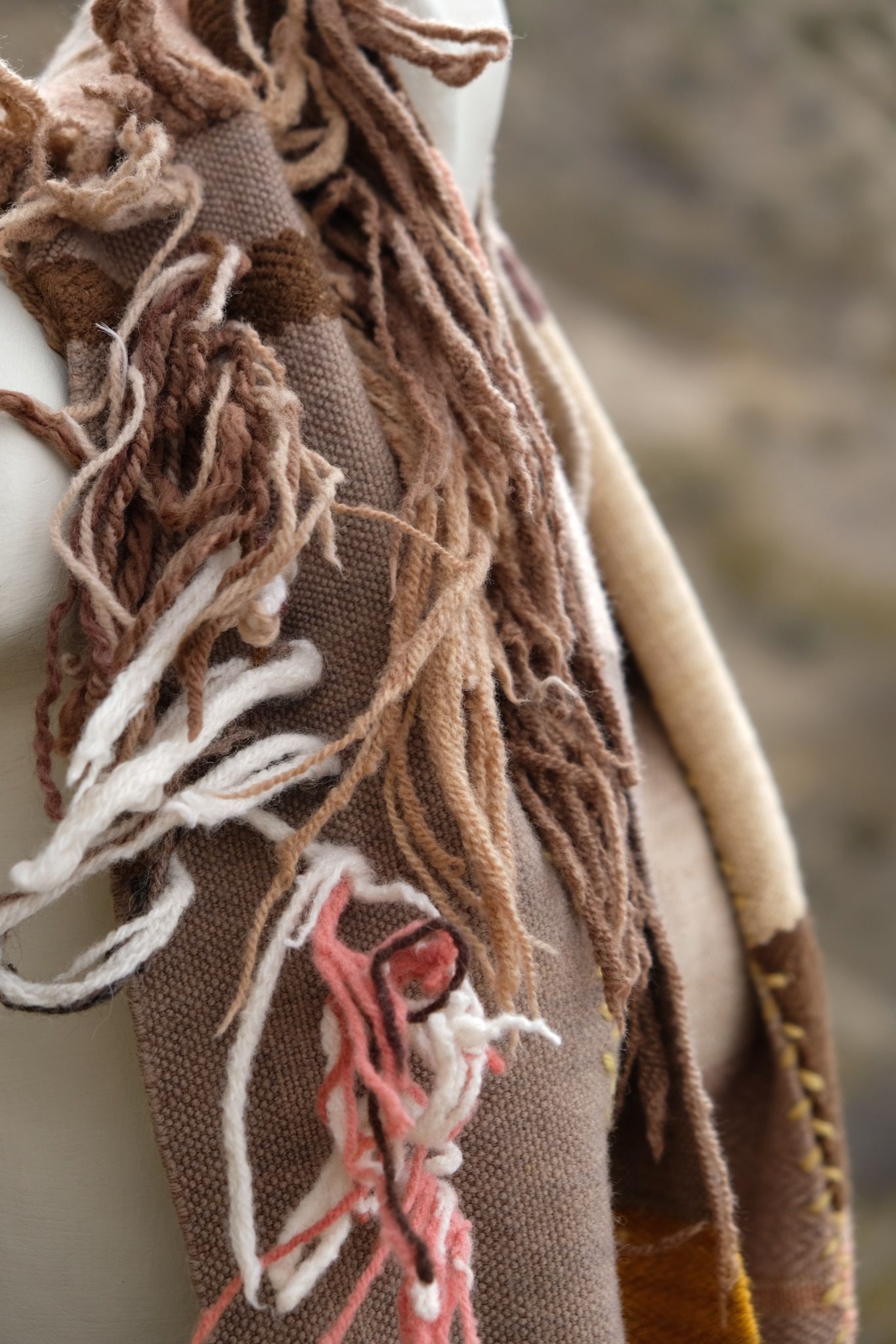 Detail of fringe on naturally dyed brown, yellow, pink, red and tan fringed infinity scarf on a white mannequin bust in the desert