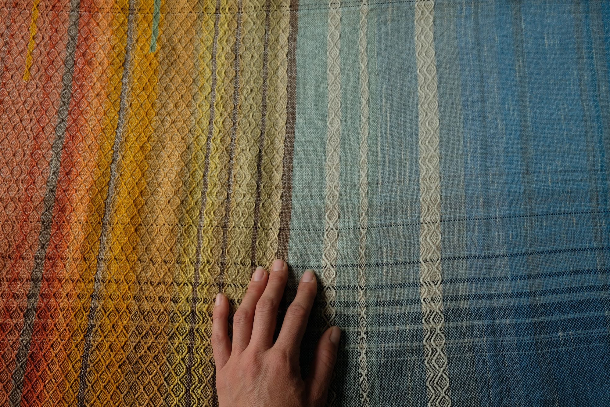 A hand resting on large brown, red, orange, yellow, blue and green piece of handwoven fabric laying on a wooden floor. 