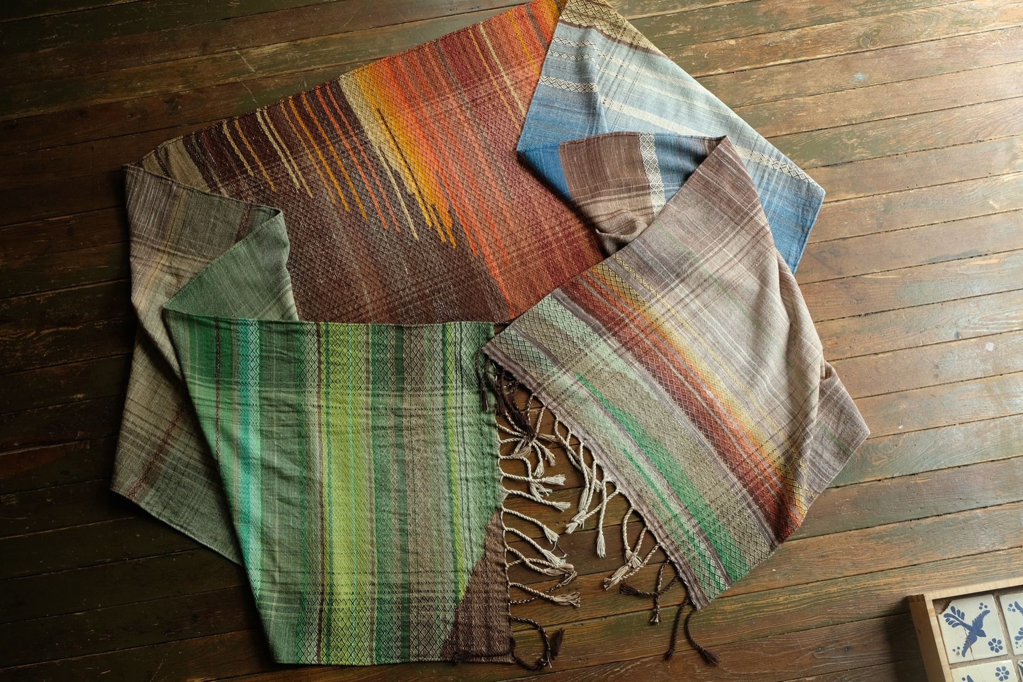 Large brown, red, orange, yellow, blue and green piece of handwoven fabric laying on a wooden floor. 