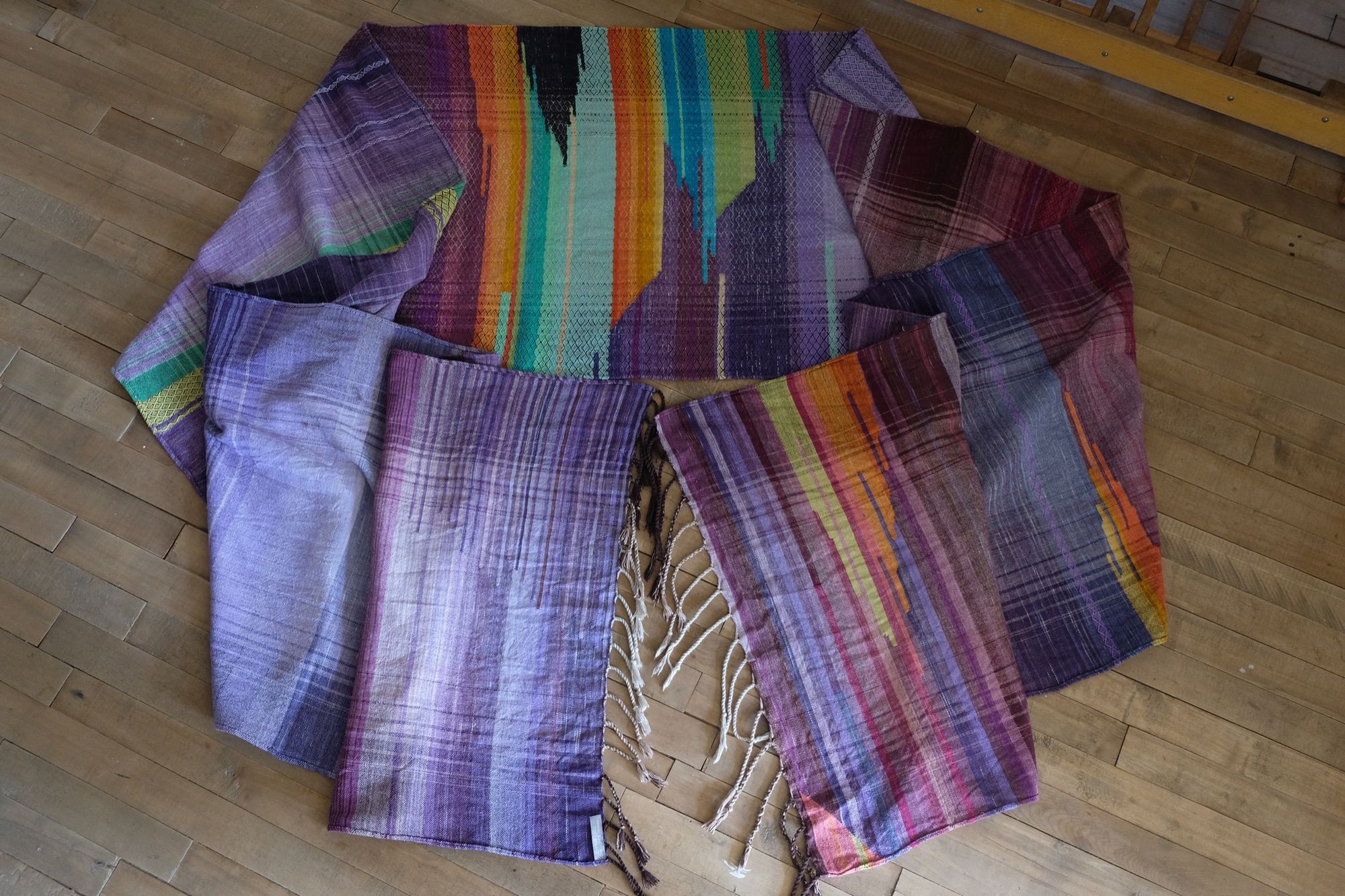 Brightly colored purple, blue, orange, green, black and pink handwoven fabric laying on a wooden floor. 