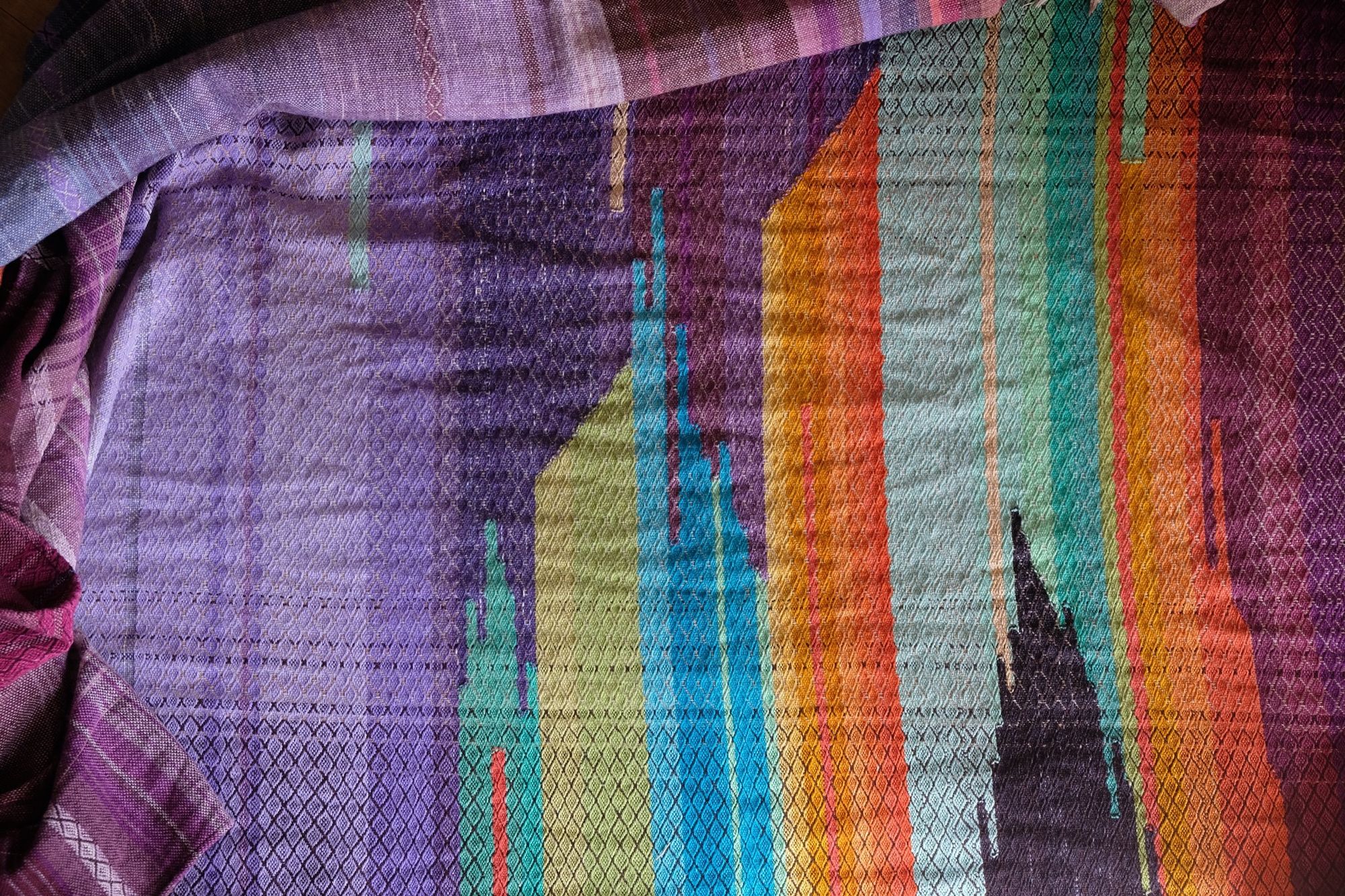 Brightly colored purple, blue, orange, green, black and pink handwoven fabric laying on a wooden floor. 