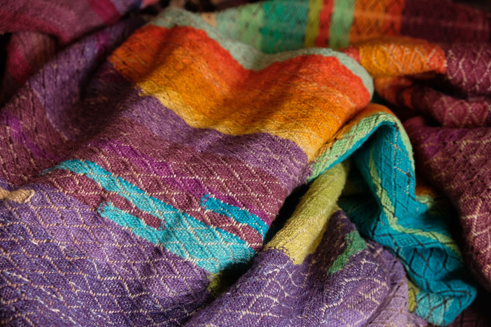 Brightly colored purple, blue, orange, green, black and pink handwoven fabric laying on a wooden floor. 