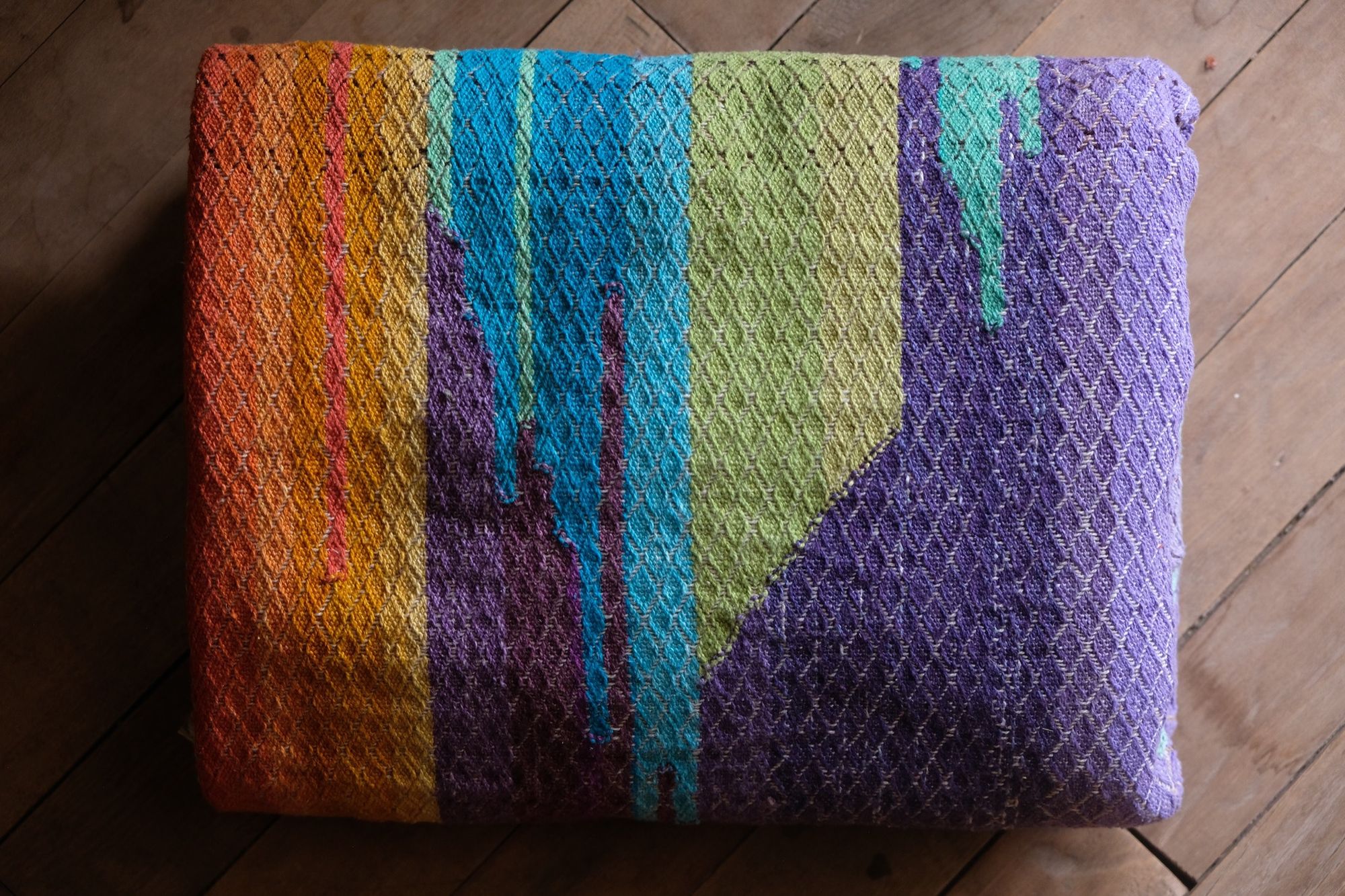 Brightly colored purple, blue, orange, green, black and pink handwoven fabric laying on a wooden floor. 