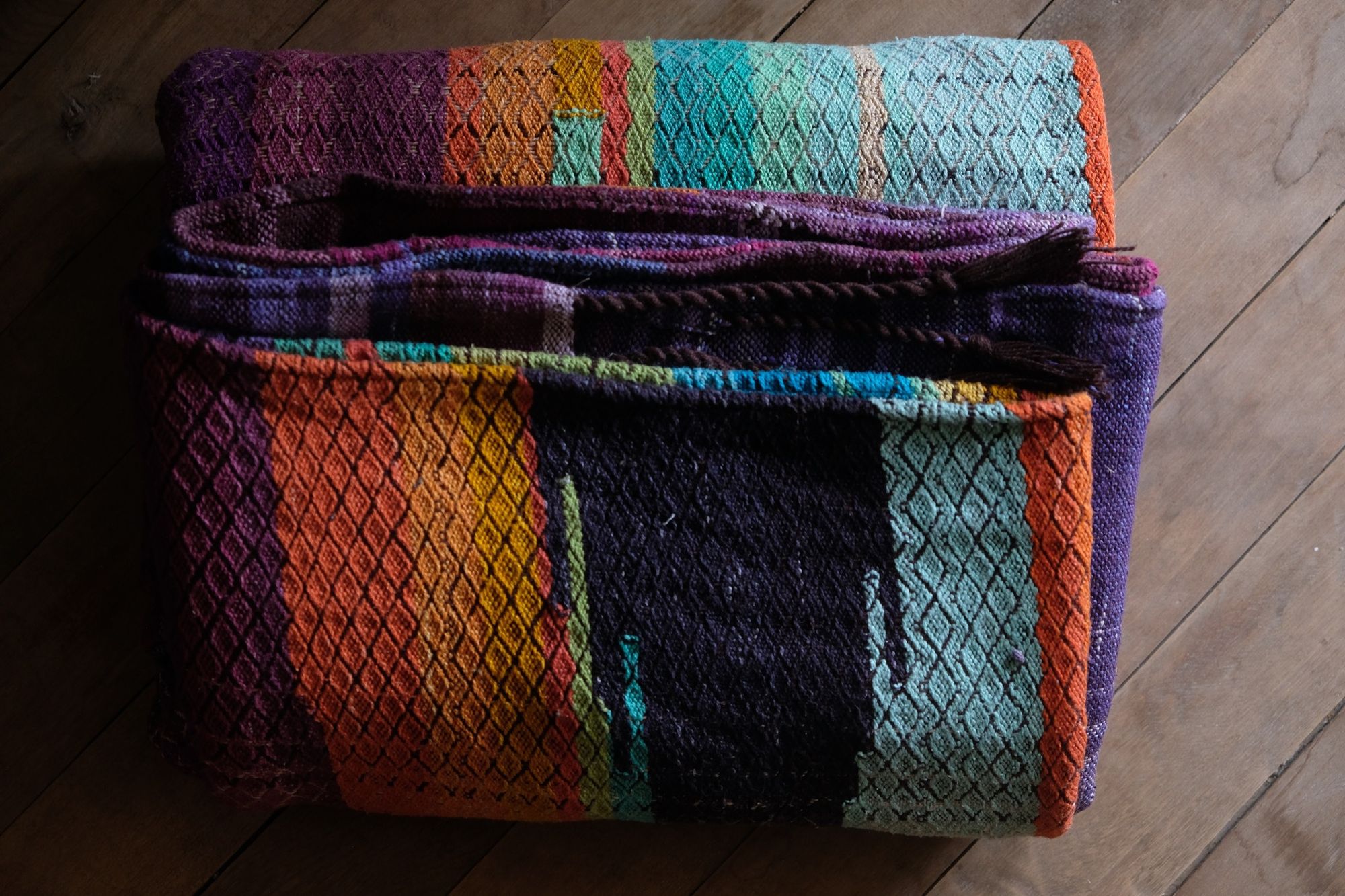 Brightly colored purple, blue, orange, green, black and pink handwoven fabric laying on a wooden floor. 