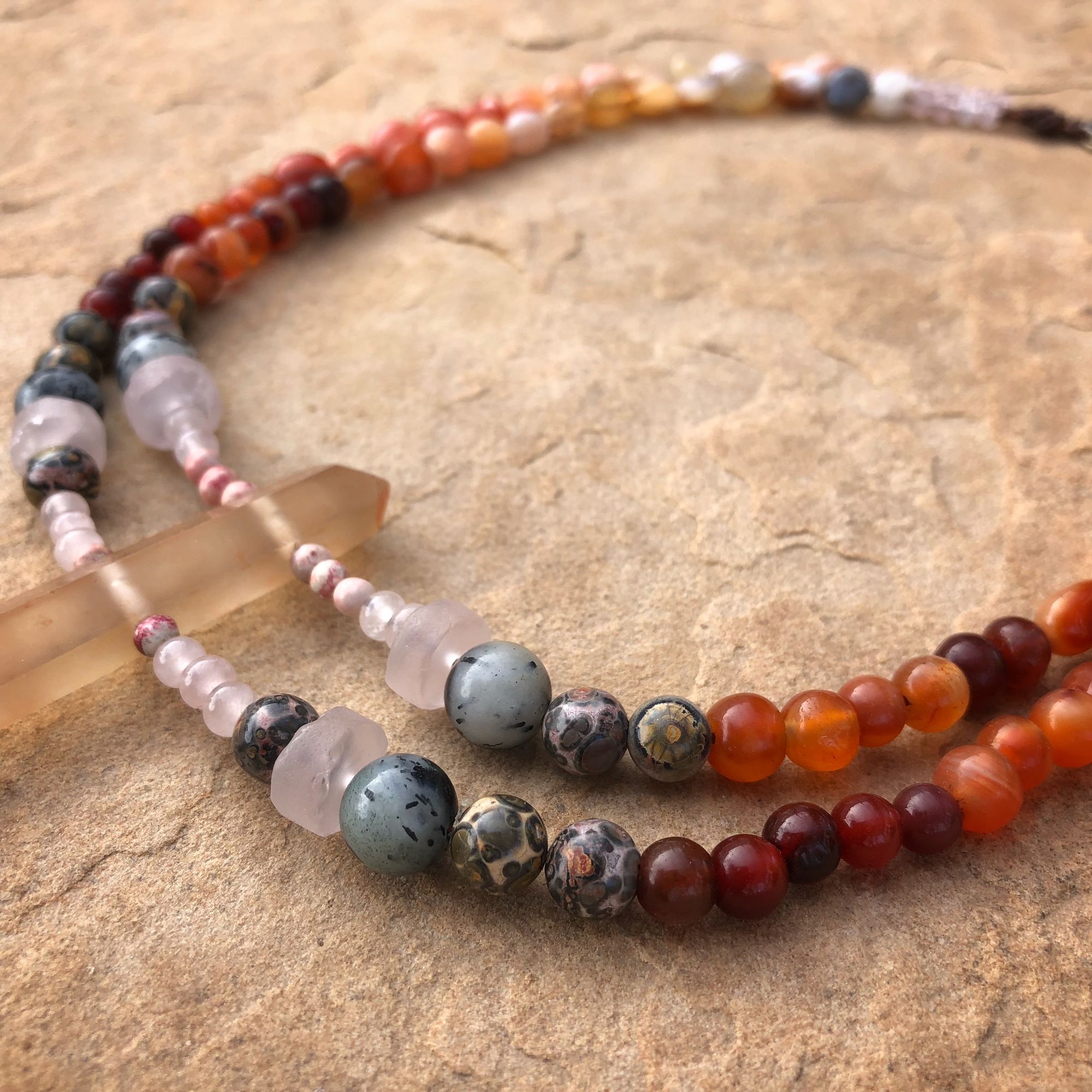 Necklace made of red, grey, orange, yellow and pink semi-precious stones laying on tan sandstone