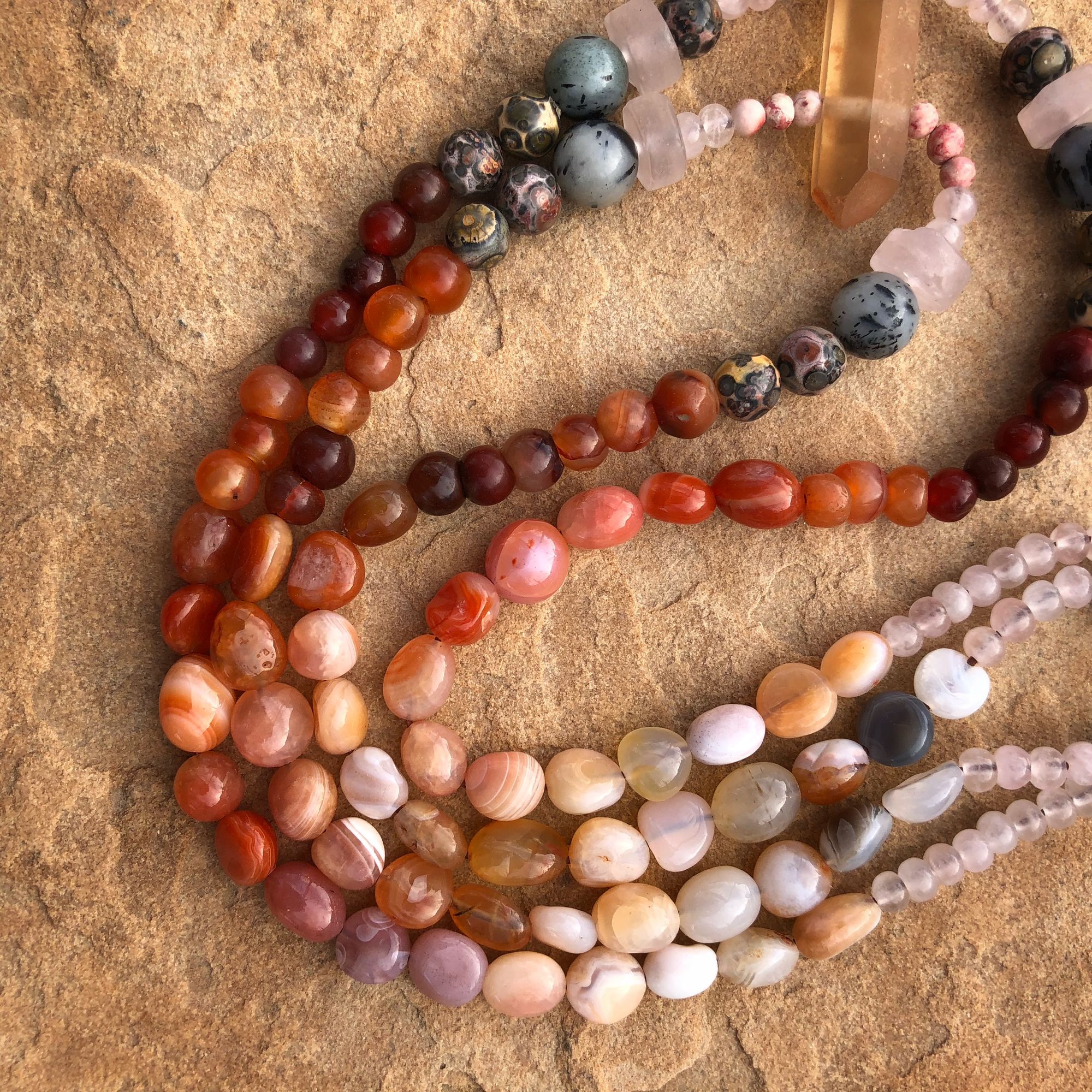 Necklace made of red, grey, orange, yellow and pink semi-precious stones laying on tan sandstone