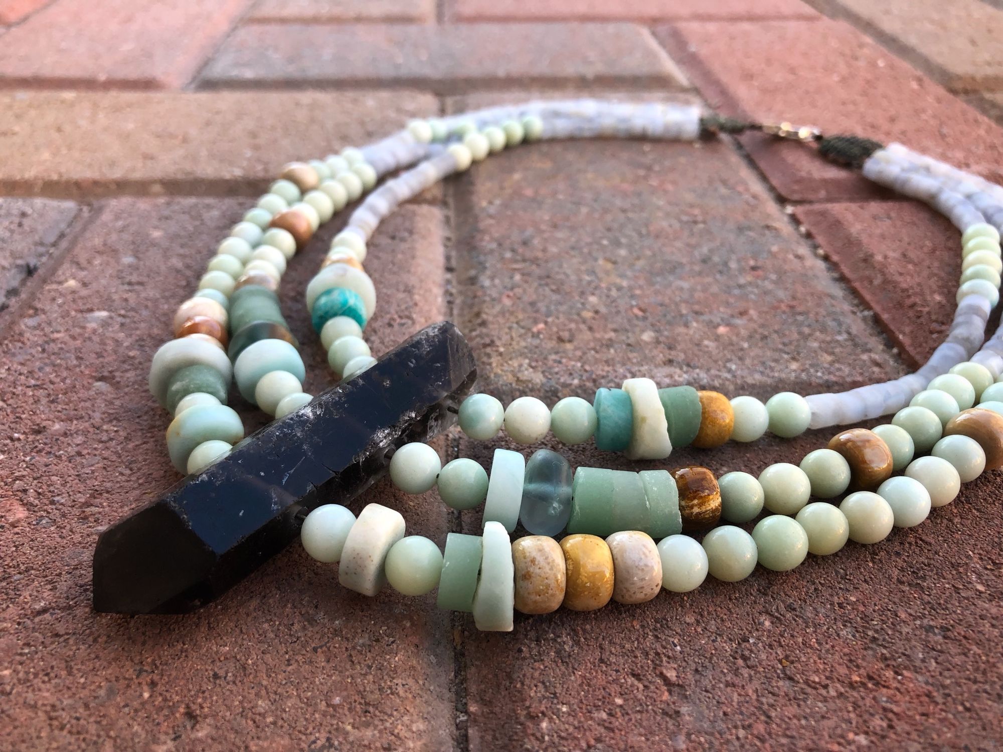 three strand necklace made with a grey crystal point and blue-green beads laying on a red brick floor