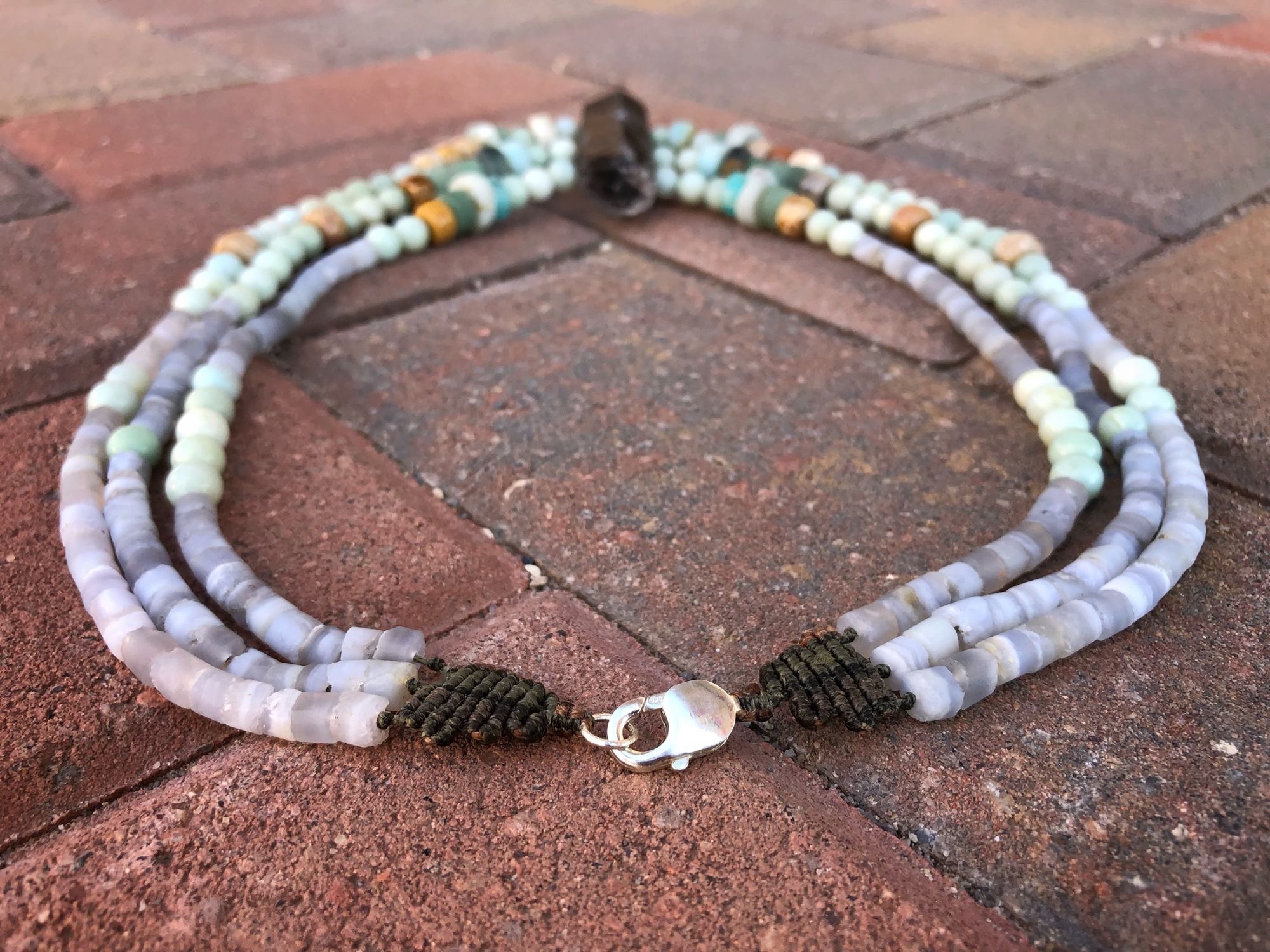 three strand necklace made with a grey crystal point and blue-green beads laying on a red brick floor