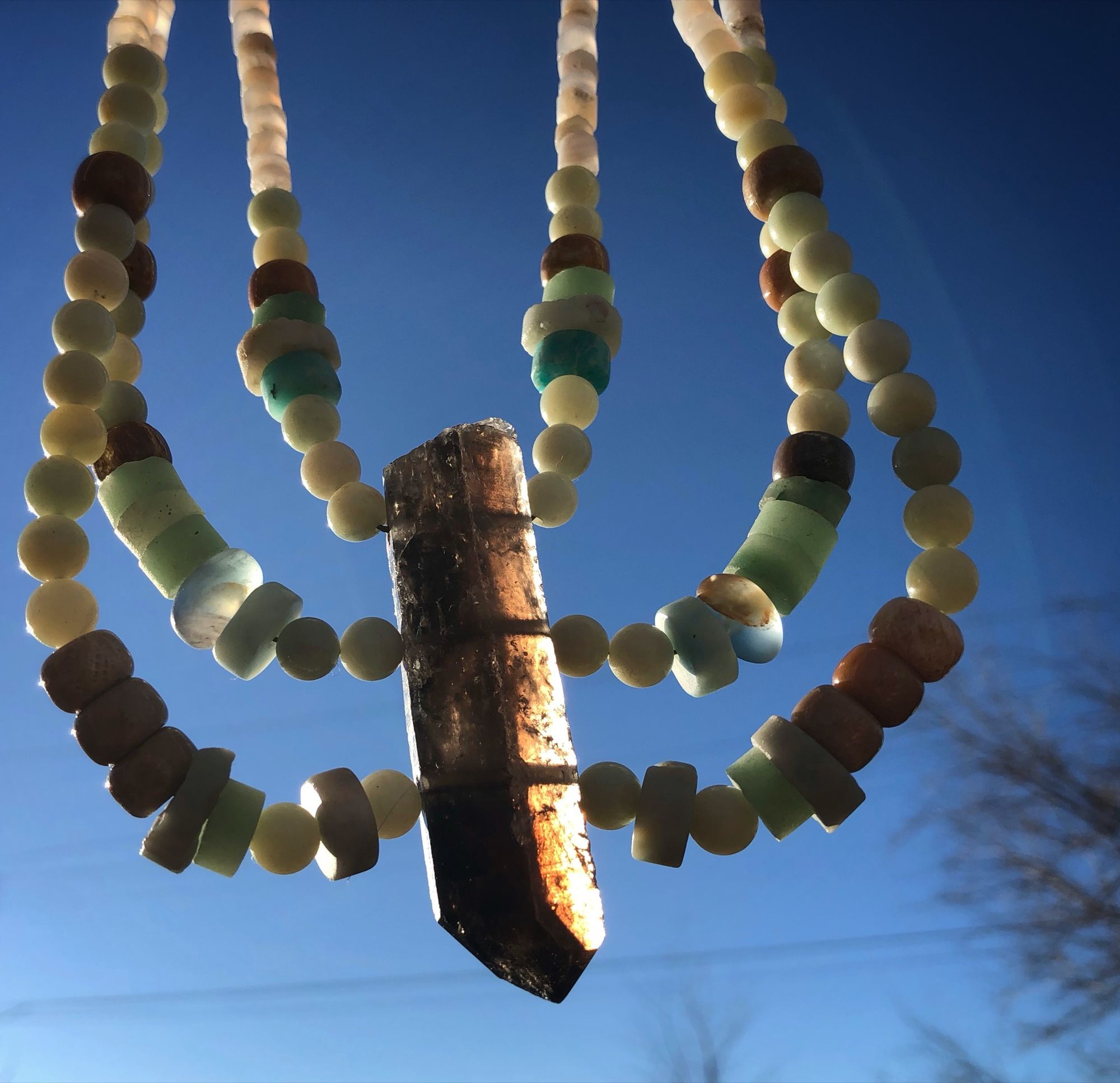 three strand necklace made with a grey crystal point and blue-green beads being held up against a blue sky