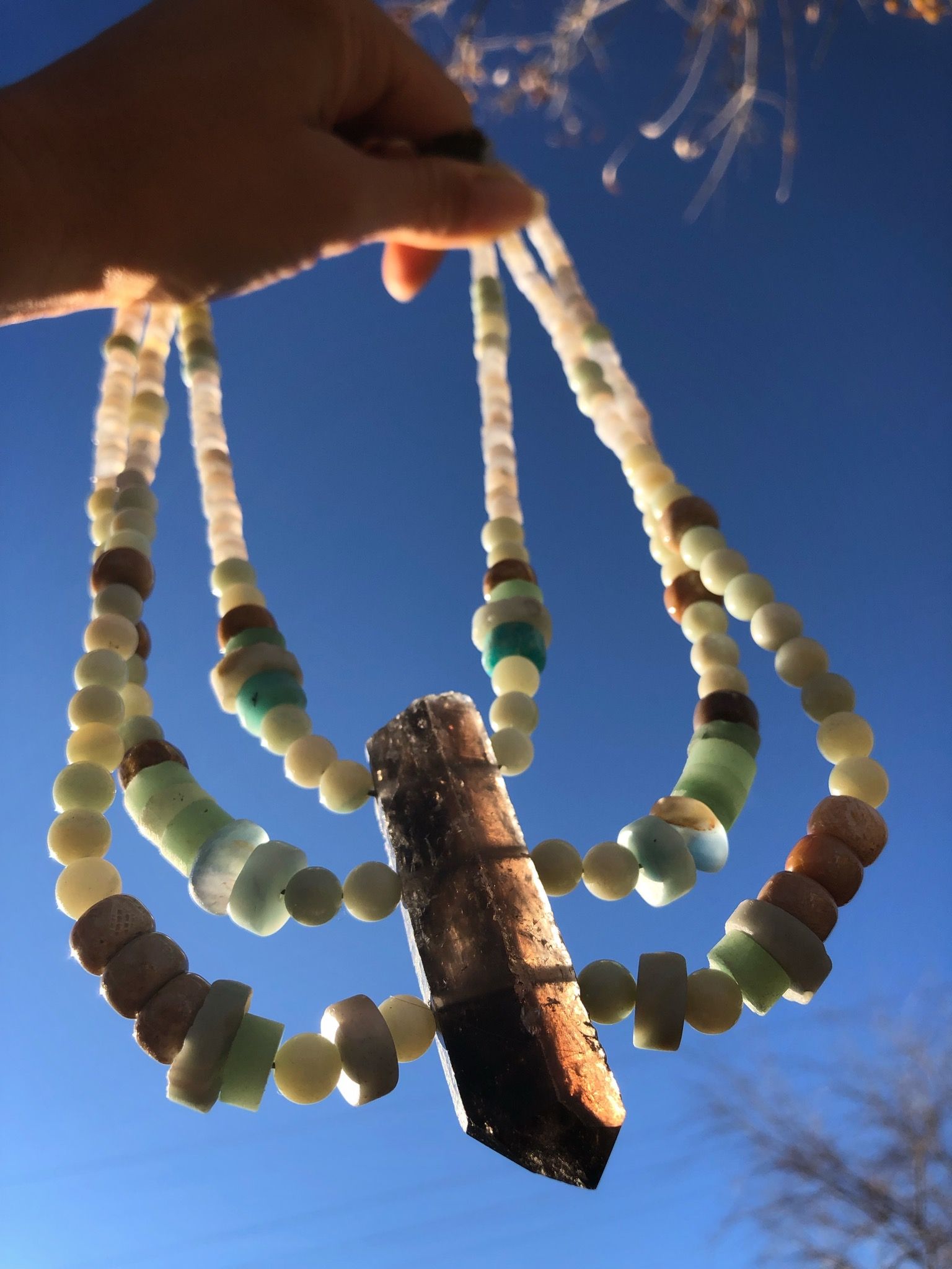 three strand necklace made with a grey crystal point and blue-green beads being held up against a blue sky