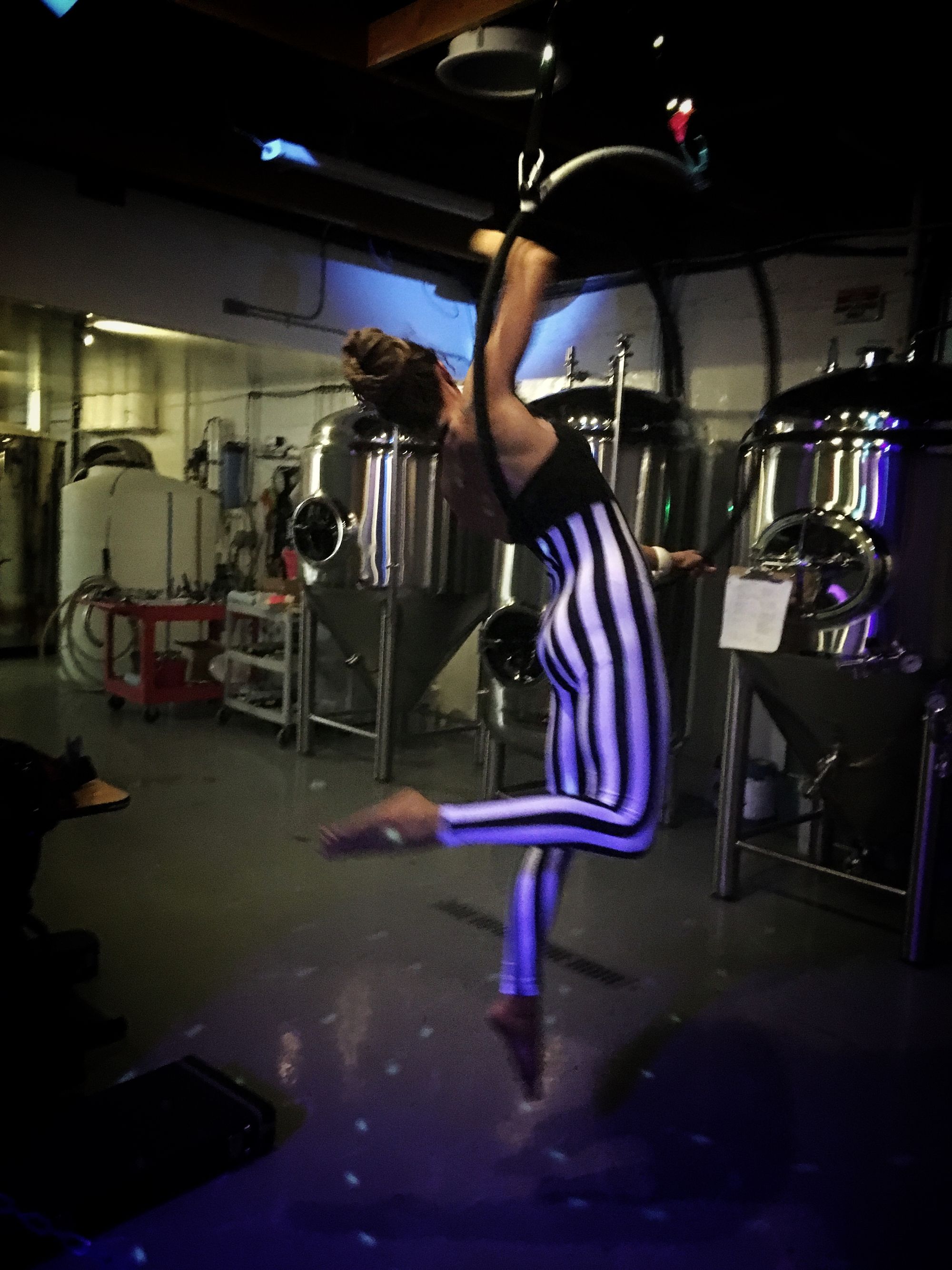 Woman performing on aerial hoop wearing black and white striped costume, next to steel tanks in a brewery