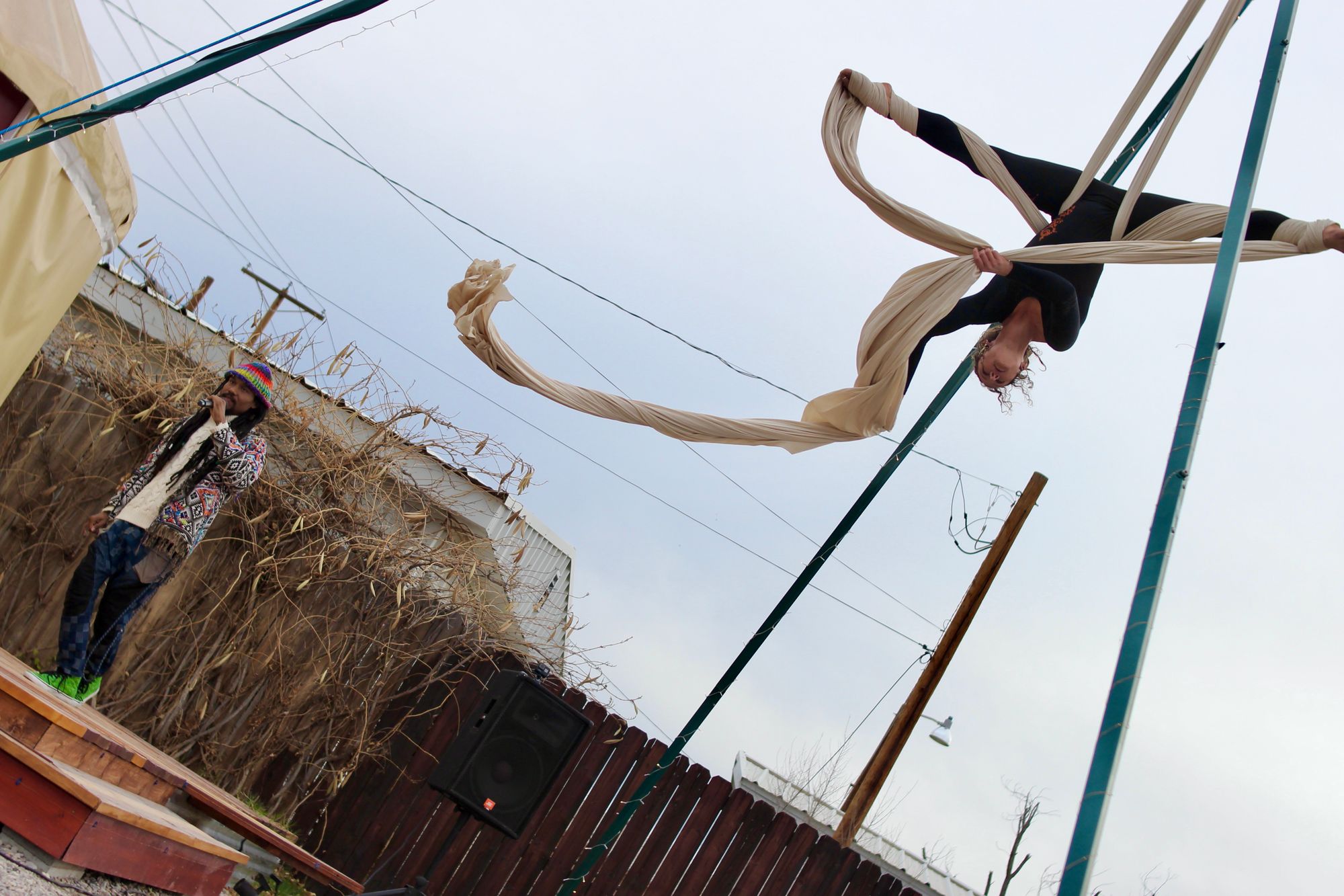 Woman performing aerial silks outside on white fabric while man holding microphone raps. 