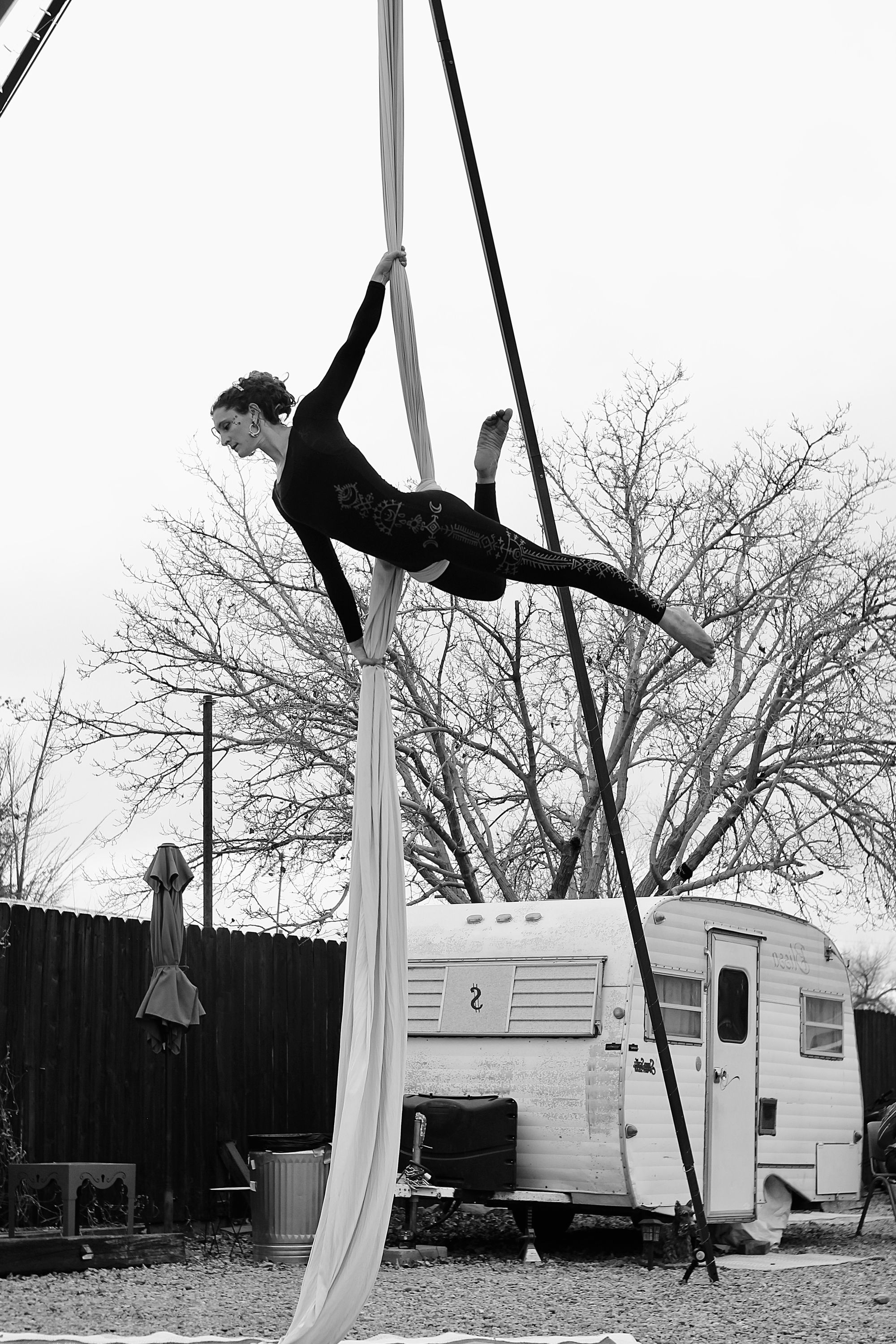 Woman performing aerial silks outside on white fabric while in front of small vintage camper trailer 
