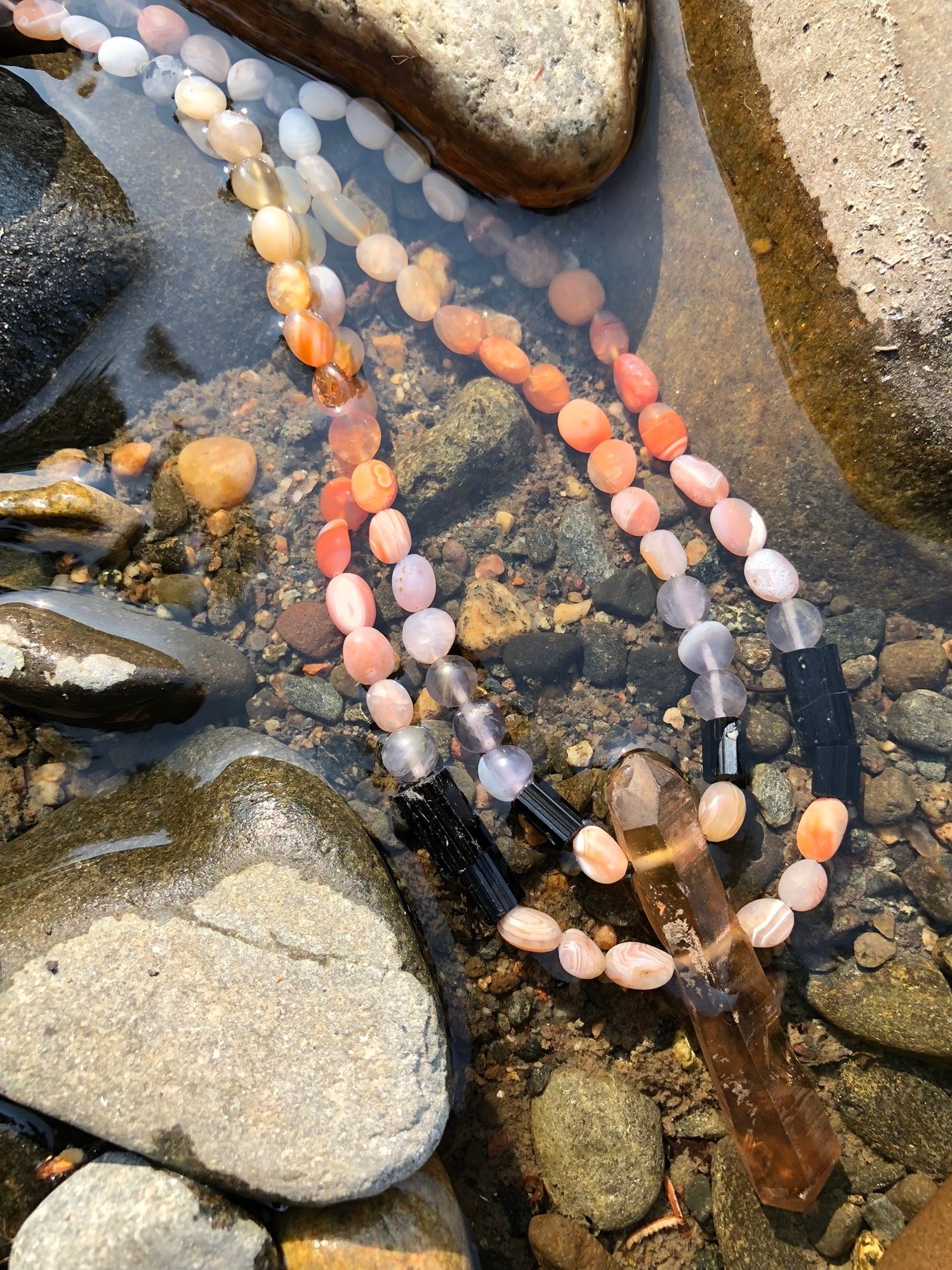 Detail of Zambian Citrine, red-orange Botswanan agate, black tourmaline, purple fluorite and larvikite necklace laying in the water of a creek