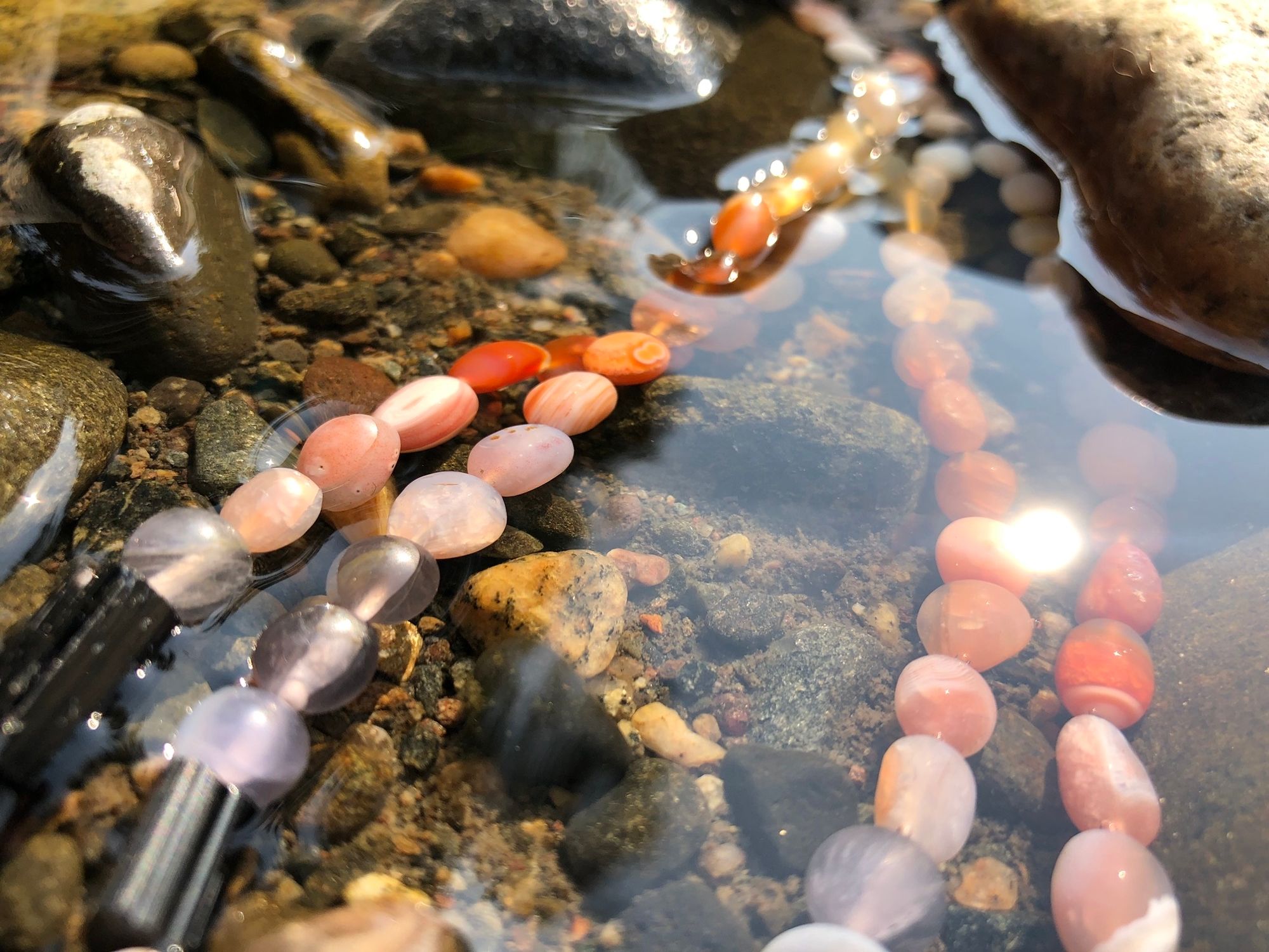 Detail of Zambian Citrine, red-orange Botswanan agate, black tourmaline, purple fluorite and larvikite necklace laying in the water of a creek
