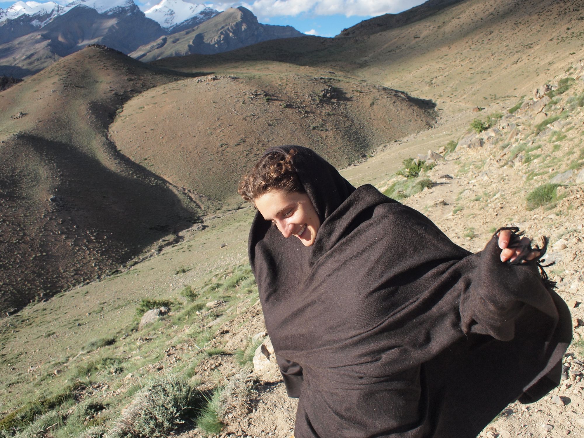 Woman in a black shawl walking on a trail in the high mountains with snow-caped peaks in the background. 