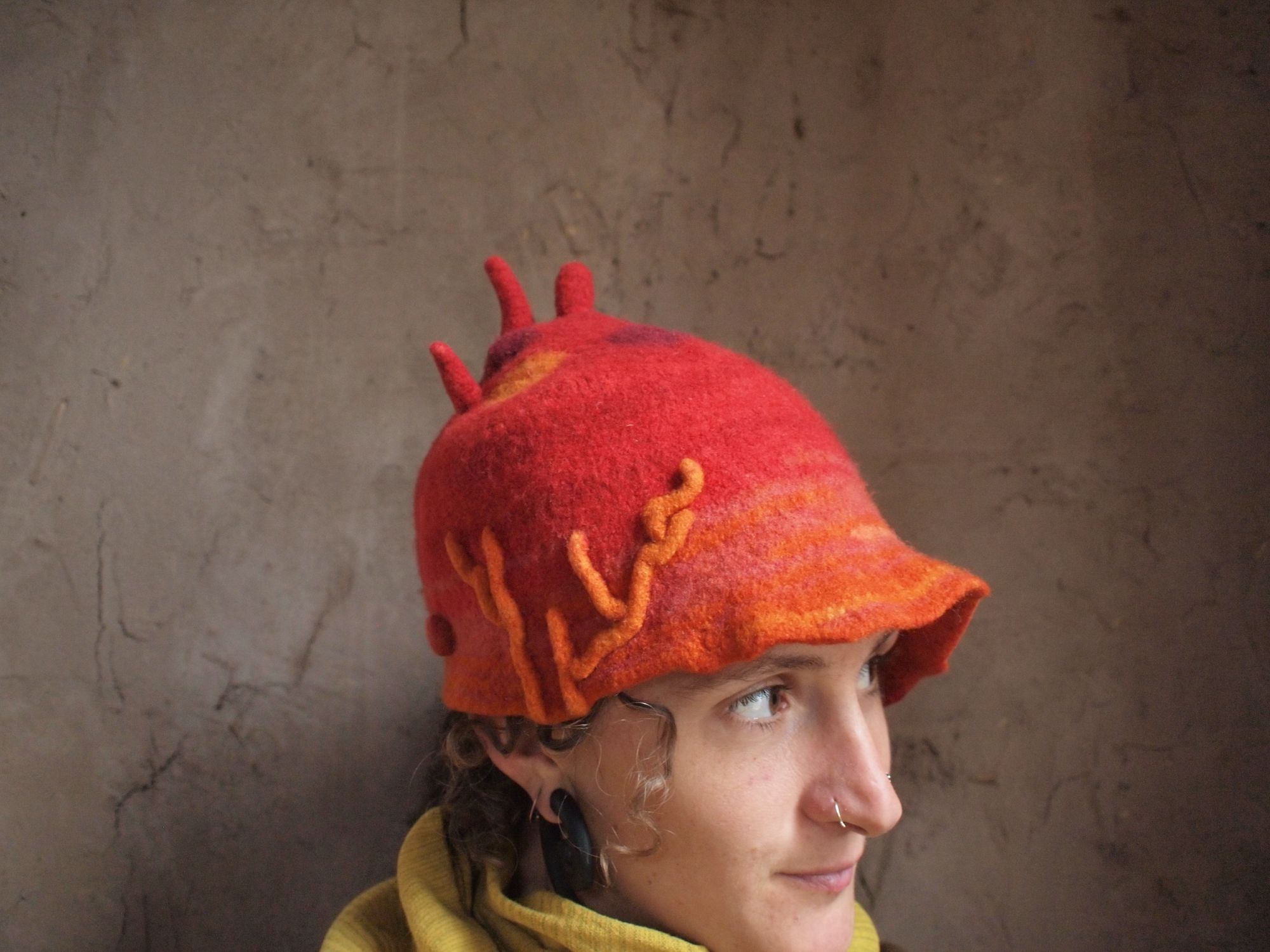 Woman wearing red and orange sculptural hat against a mud wall. 