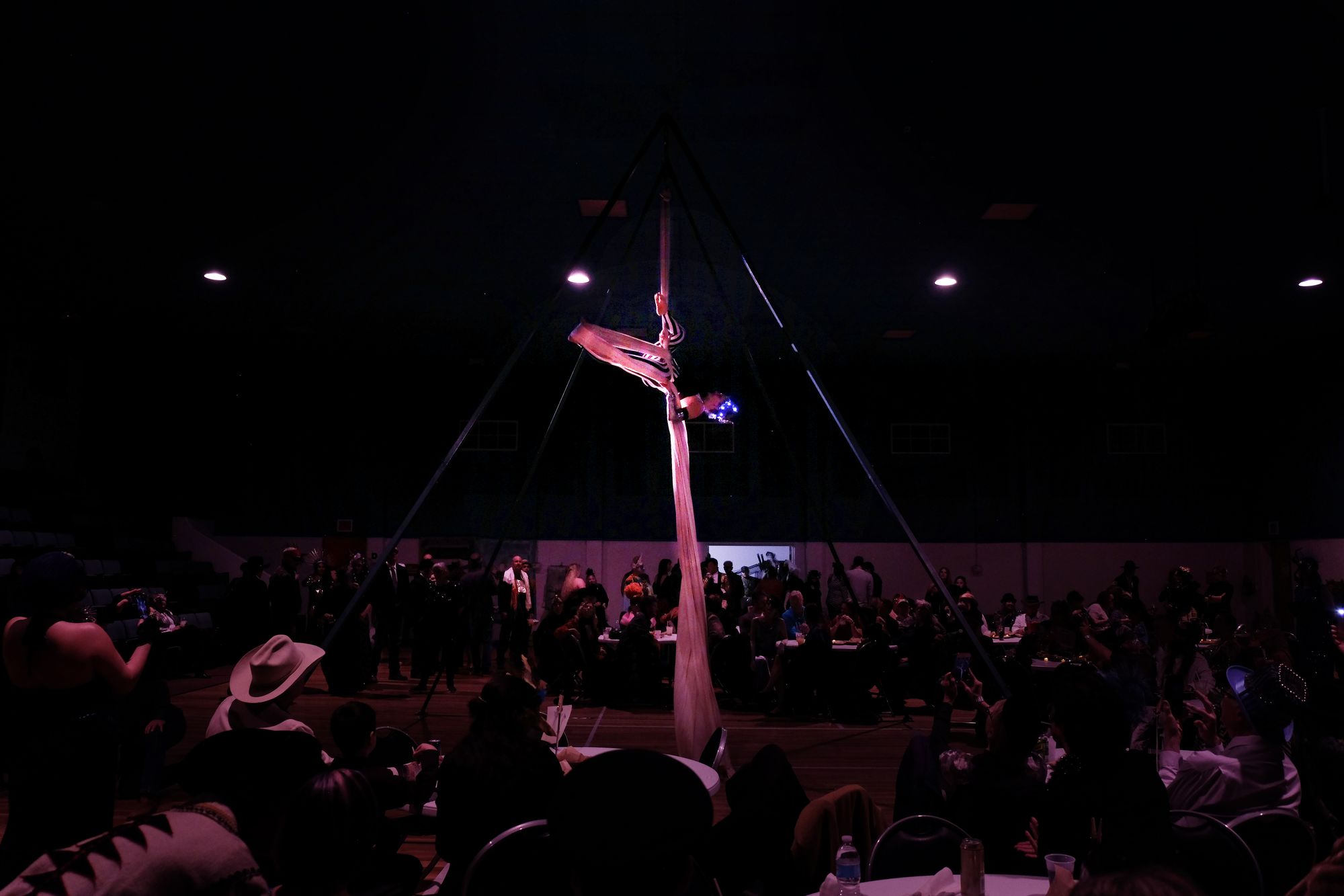 Woman performing aerial silks on white fabric in a dimly lit gymnasium at a masquerade ball