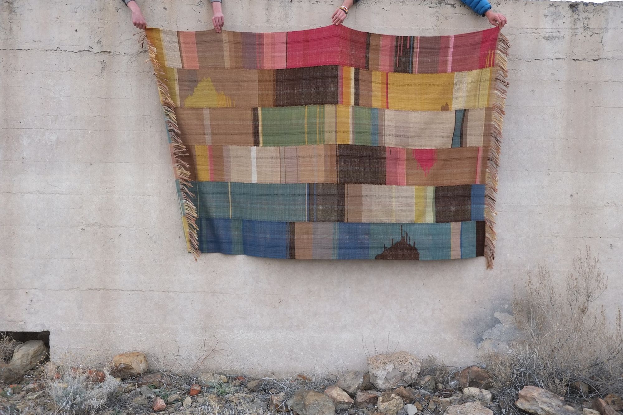 A multicolored woolen blanket being held by four hands agains a white concrete wall.