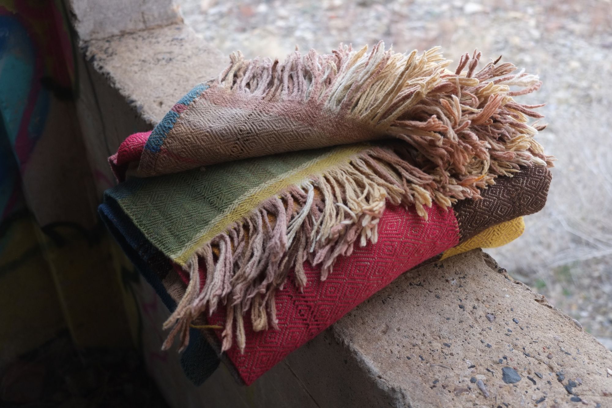 Details of a brown, pink, yellow, blue, green, tan and purple woolen blanket with a diamond pattern folded and resting on a concrete window frame .