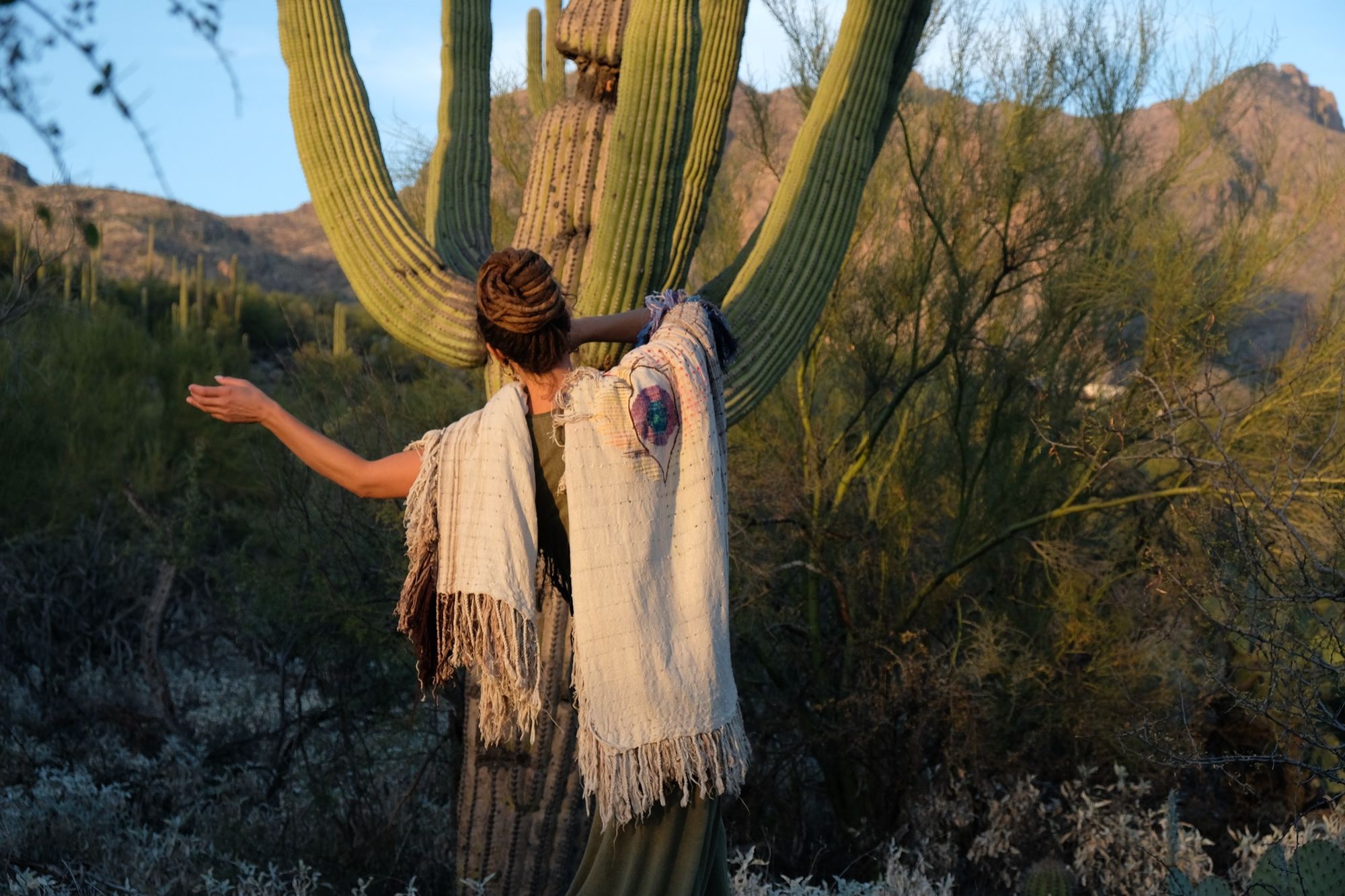Woman wearing a rainbow, white and brown colored shawl, completely covered in fringe in the desert with cactus and mountains