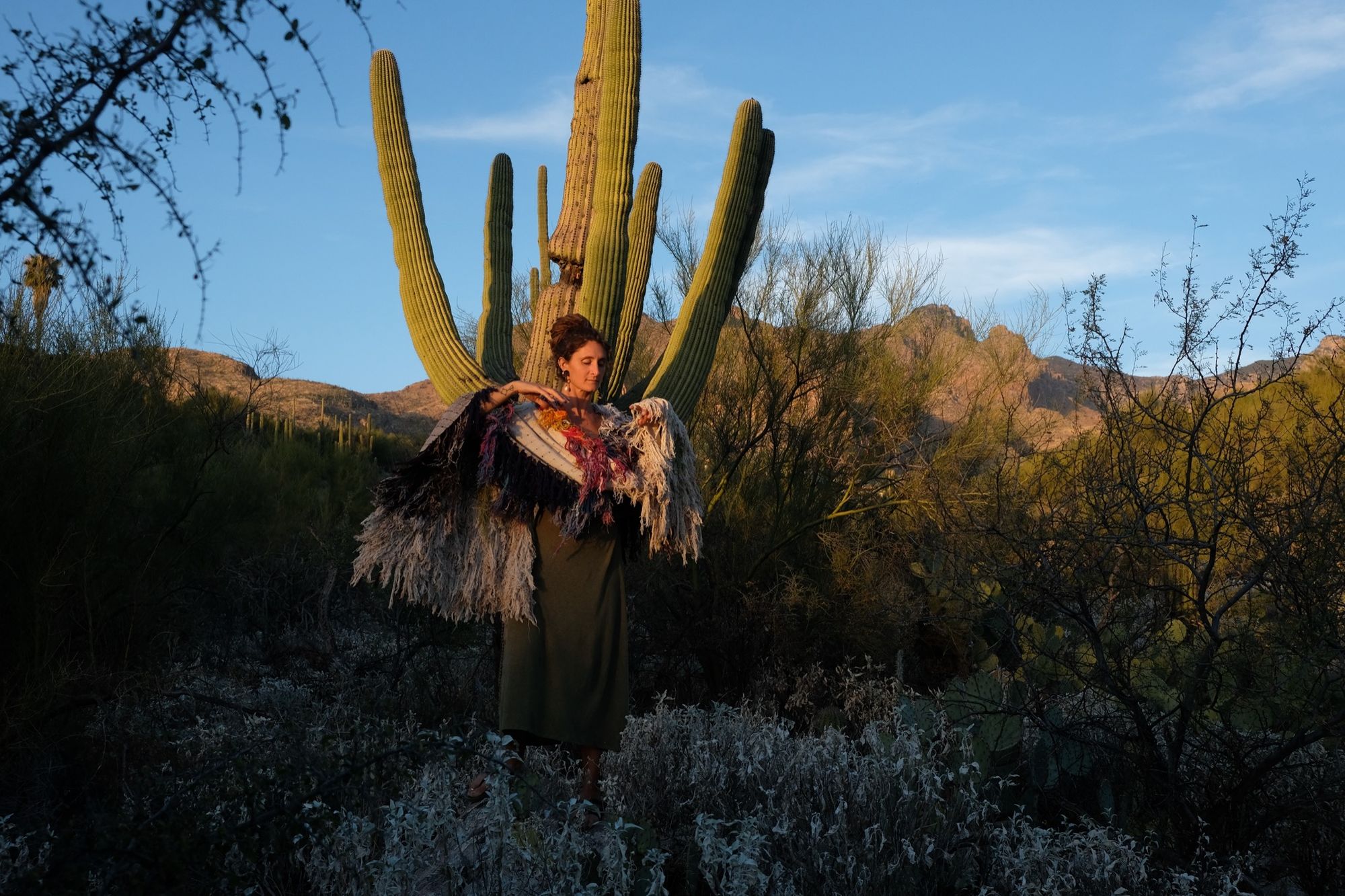Woman wearing a rainbow, white and brown colored shawl, completely covered in fringe in the desert with cactus and mountains