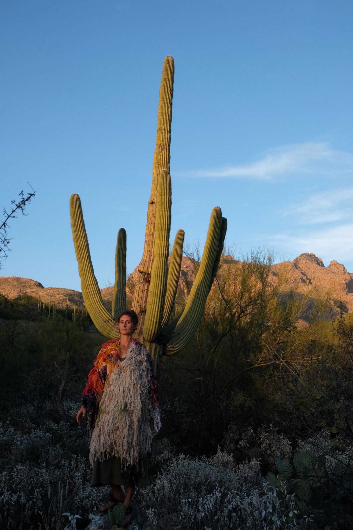 Woman wearing a rainbow, white and brown colored shawl, completely covered in fringe in the desert with cactus and mountains