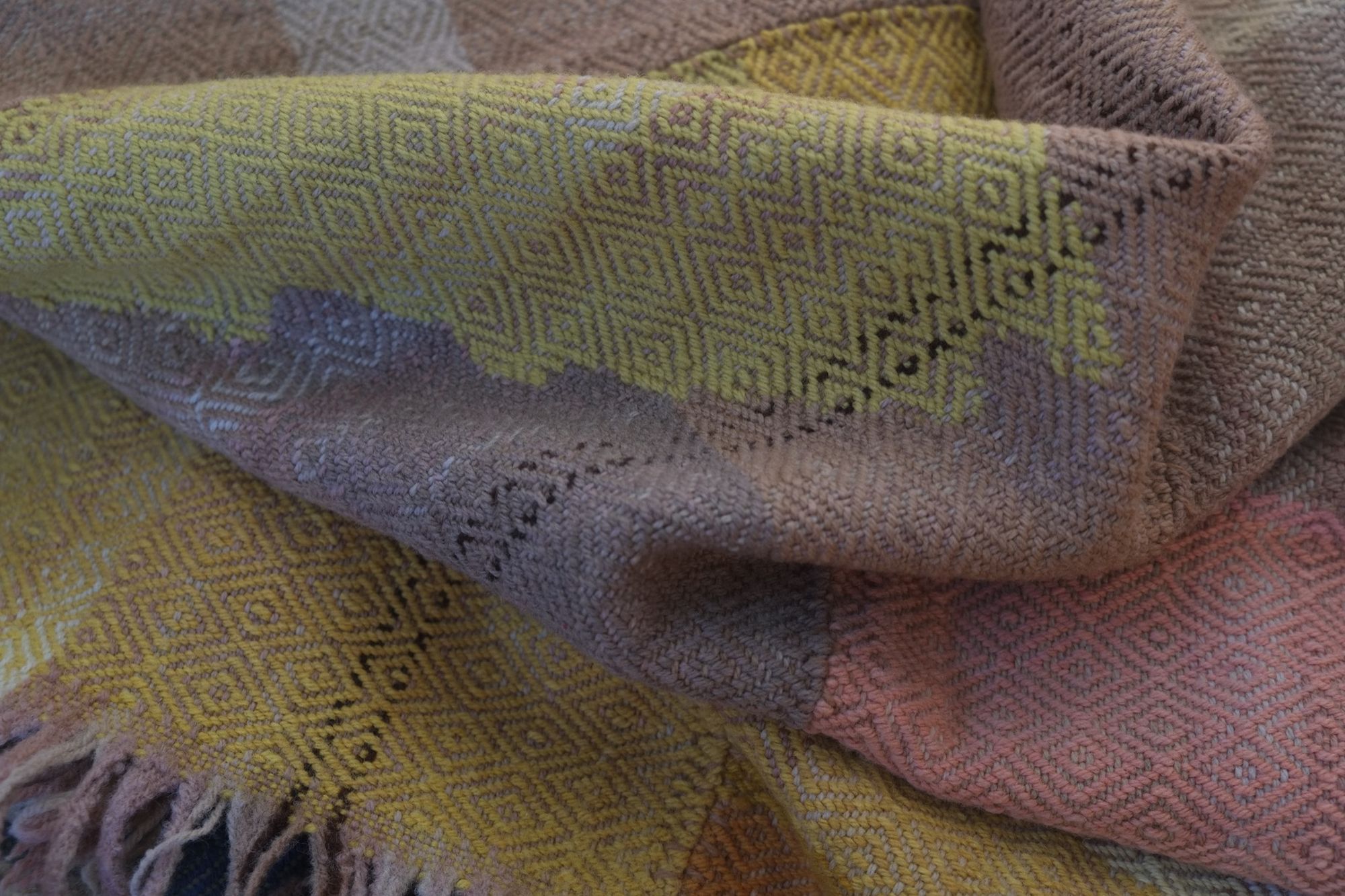 Details of a brown, pink, yellow, blue, green, tan and purple woolen blanket with a diamond pattern.