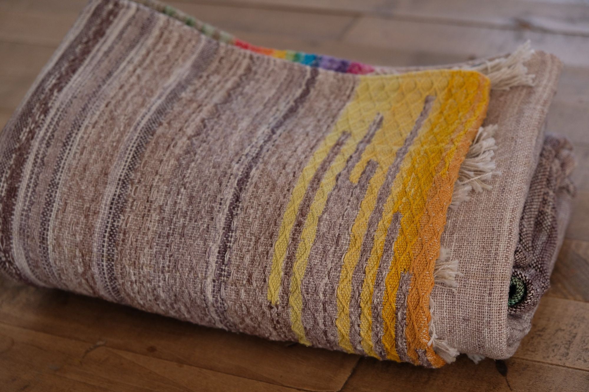 Handwoven raw silk fabric with textures diamond weave in browns, tans, purples, blues, yellows, pink and orange folded and laying on a wood floor