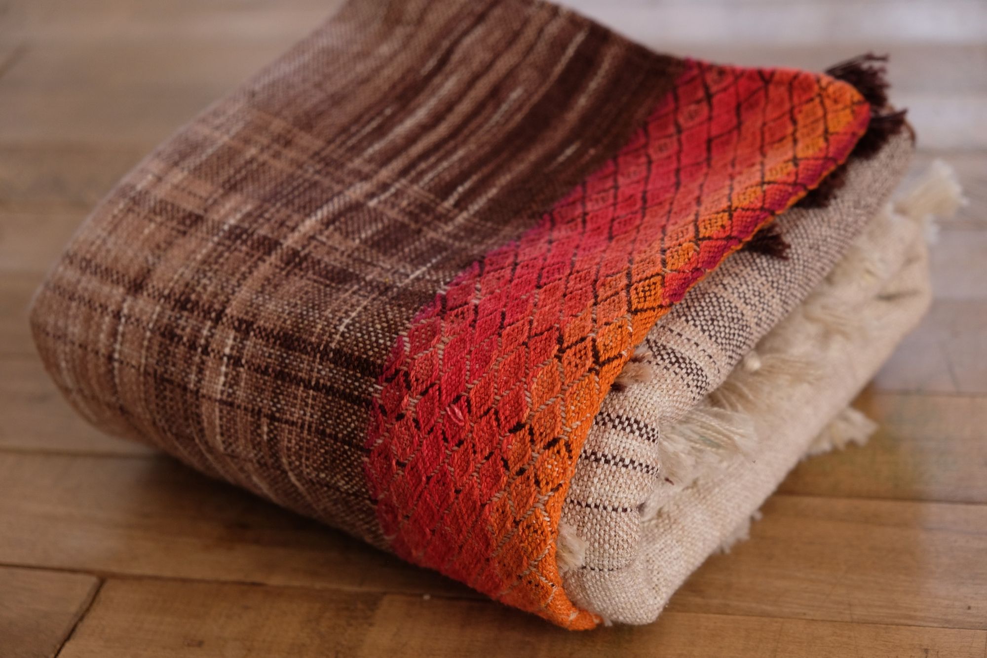detail of handwoven fabric with textured diamond weave in rainbow colors with browns and tans folded on a wooden floor