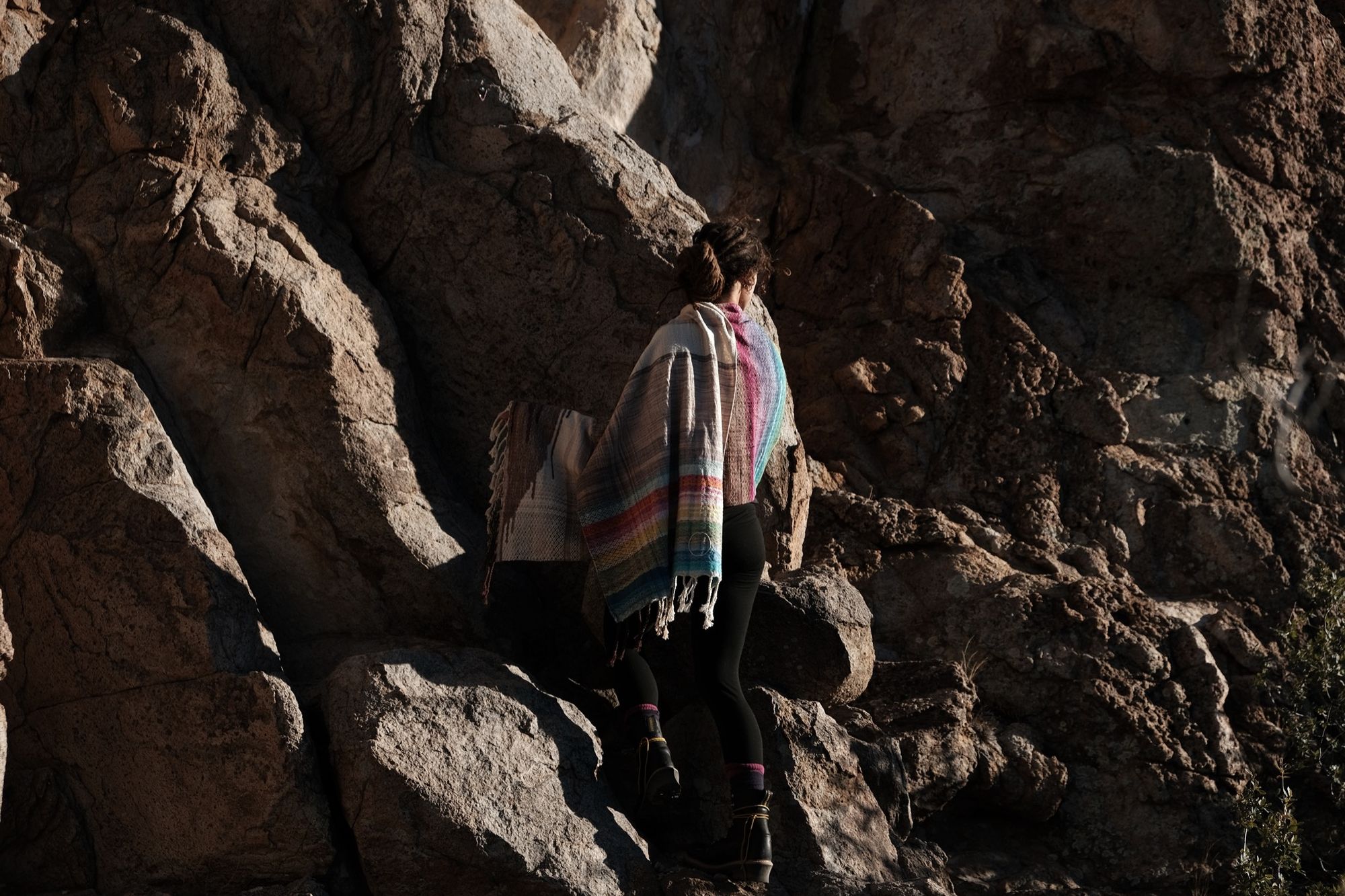 woman wearing all black and a rainbow colored scarf shawl, sitting on rocks at the base of a cliff