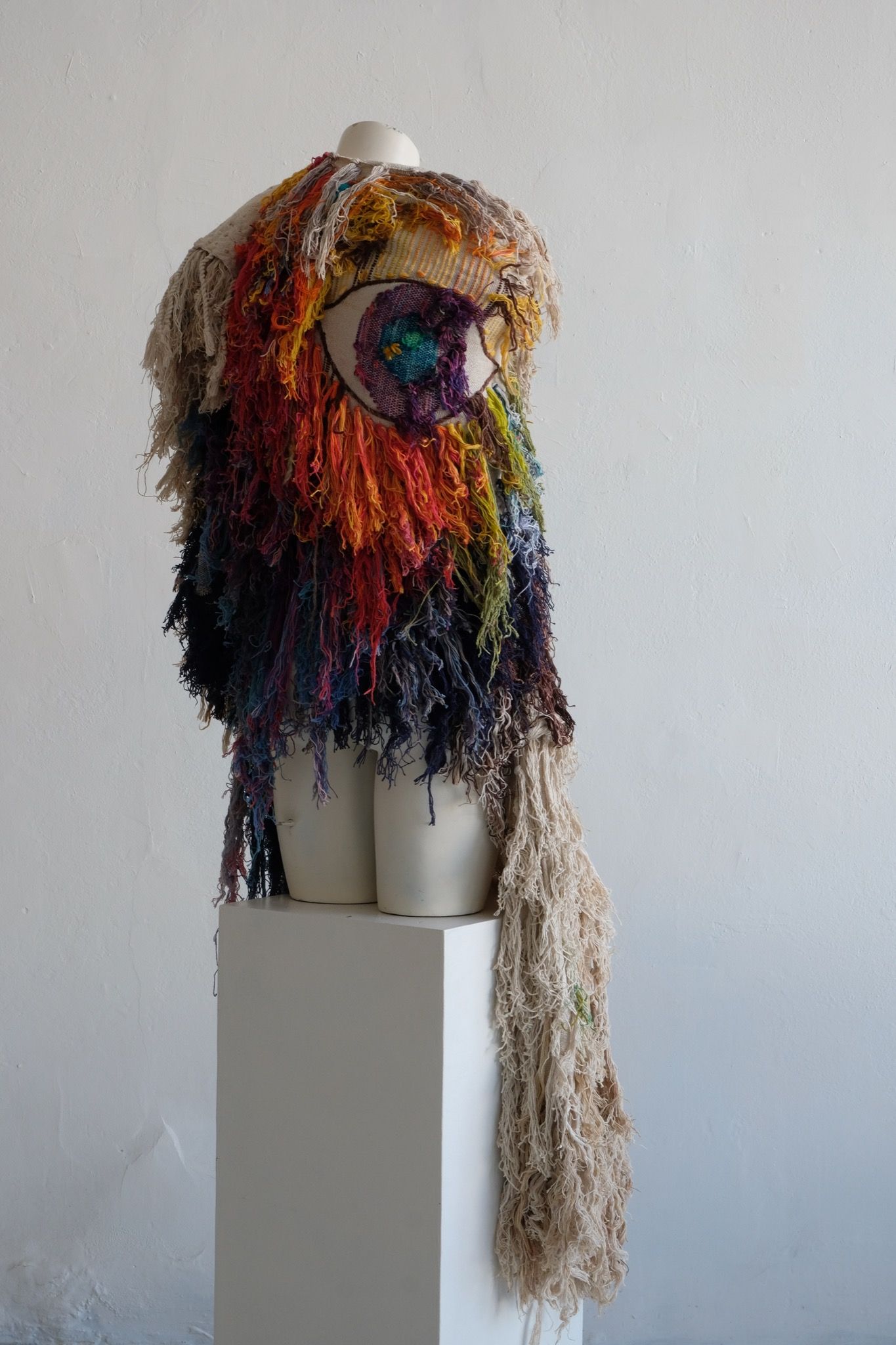 A rainbow, white and brown colored shawl, completely covered in fringe on a white mannequin, on a white pedestal, in a white room