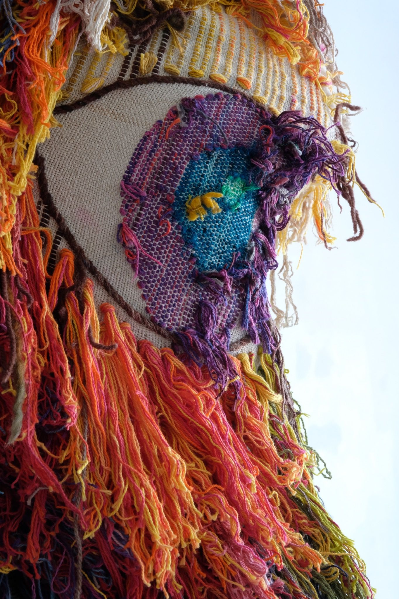 detail of A rainbow, white and brown colored shawl, completely covered in fringe on a white mannequin, on a white pedestal, in a white room