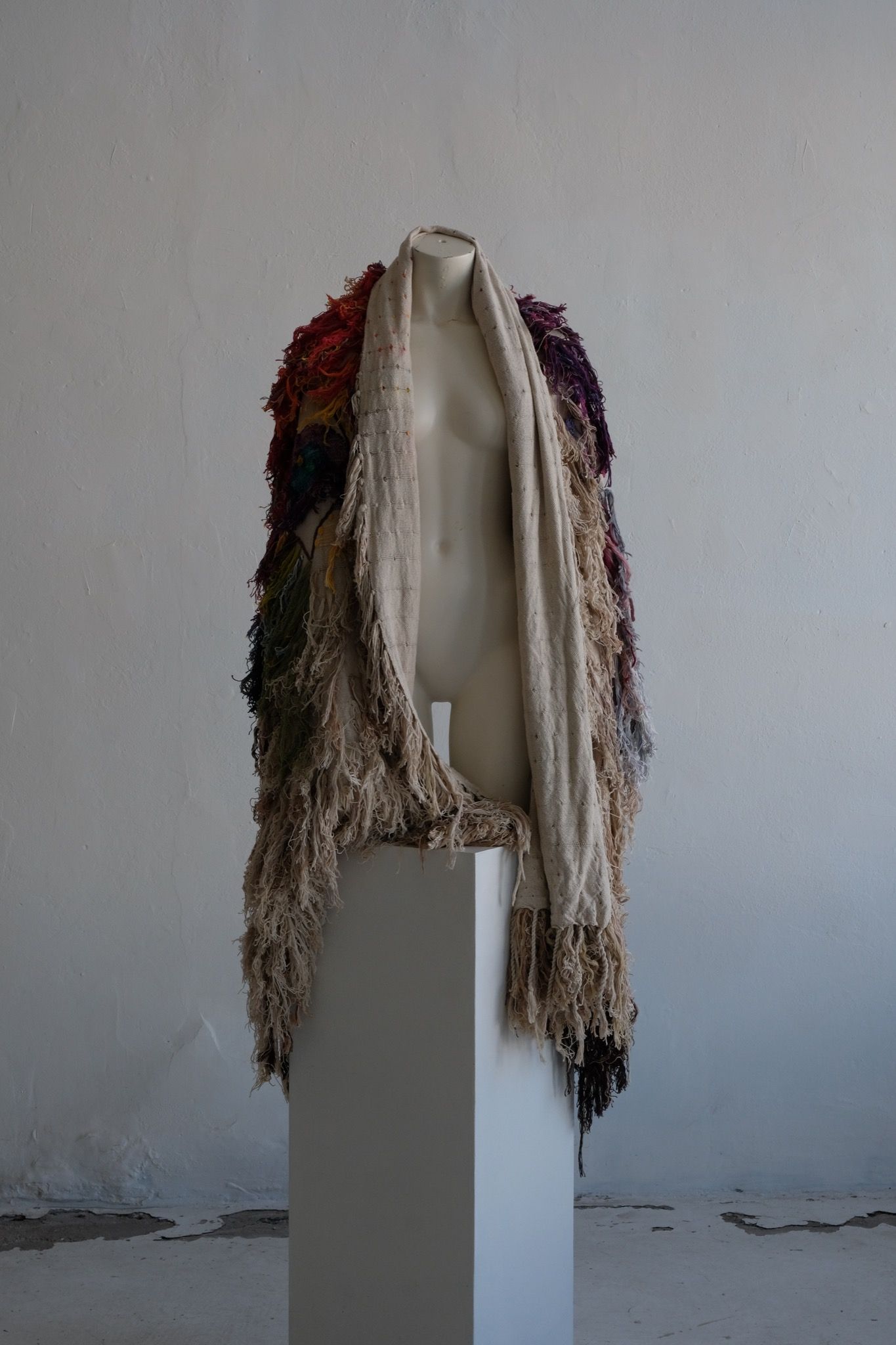 A rainbow, white and brown colored shawl, completely covered in fringe on a white mannequin, on a white pedestal, in a white room