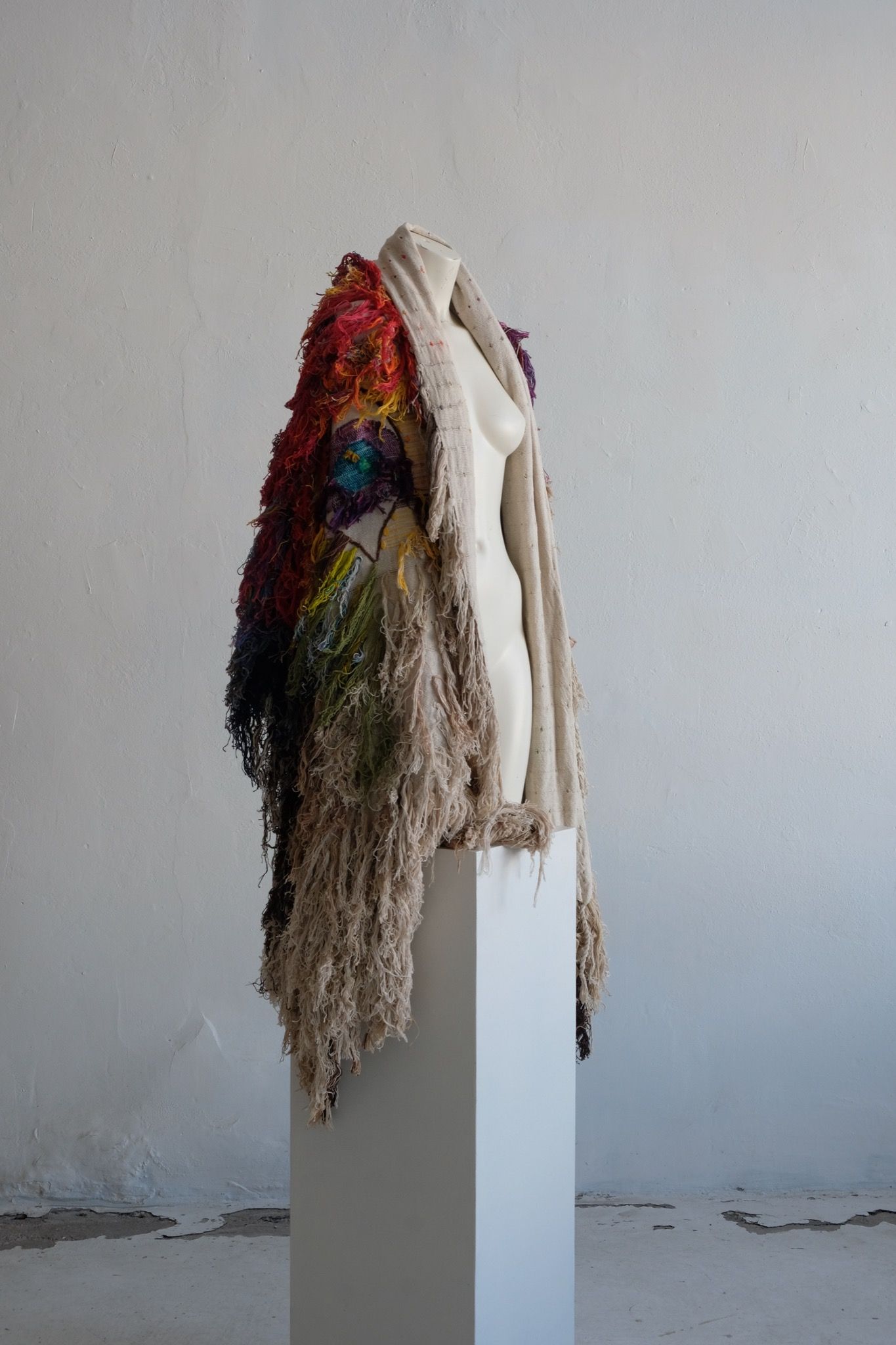 A rainbow, white and brown colored shawl, completely covered in fringe on a white mannequin, on a white pedestal, in a white room