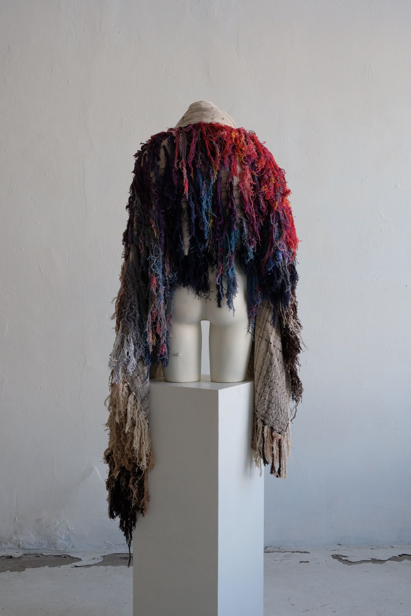 A rainbow, white and brown colored shawl, completely covered in fringe on a white mannequin, on a white pedestal, in a white room