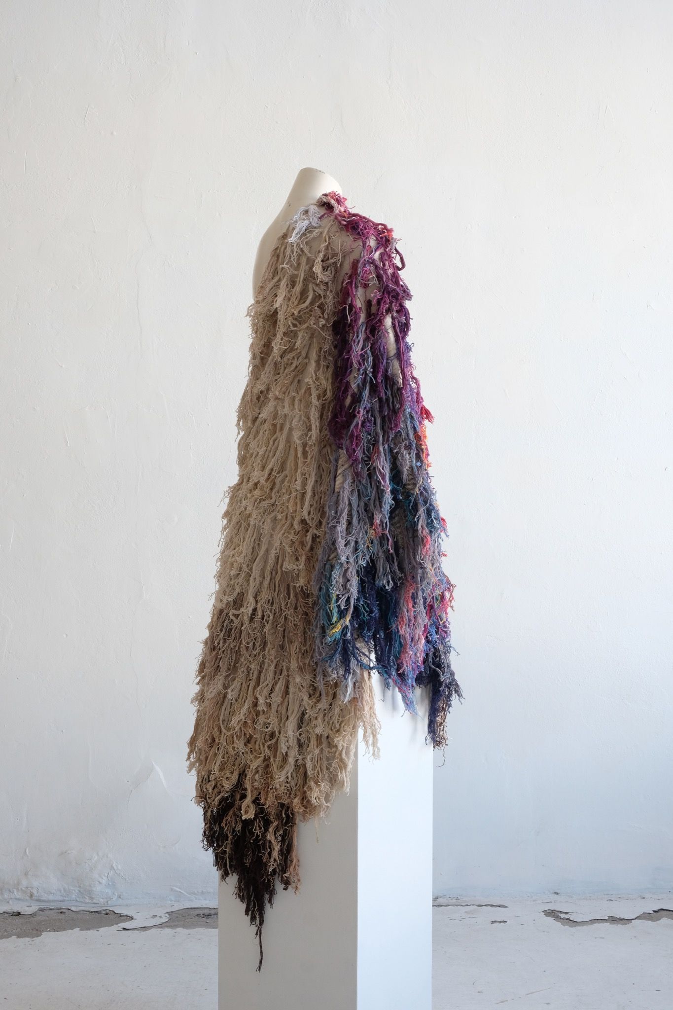 A rainbow, white and brown colored shawl, completely covered in fringe on a white mannequin, on a white pedestal, in a white room