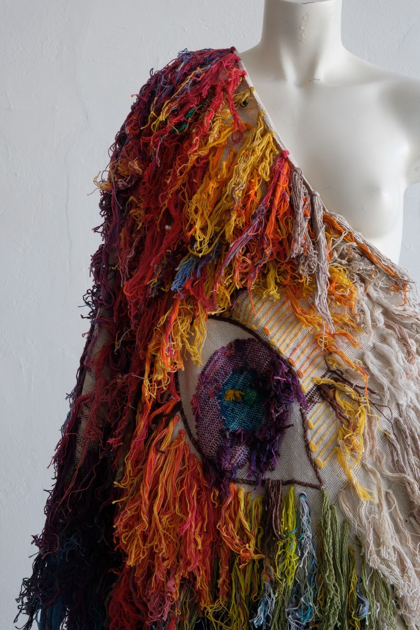 A rainbow, white and brown colored shawl, completely covered in fringe on a white mannequin, on a white pedestal, in a white room