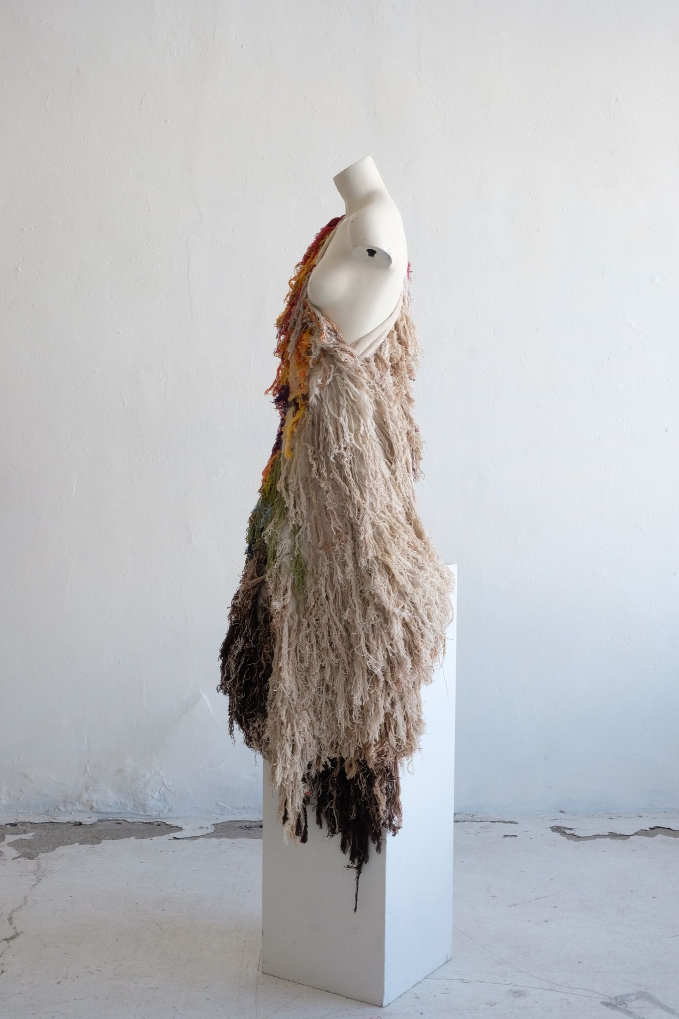 A rainbow, white and brown colored shawl, completely covered in fringe on a white mannequin, on a white pedestal, in a white room