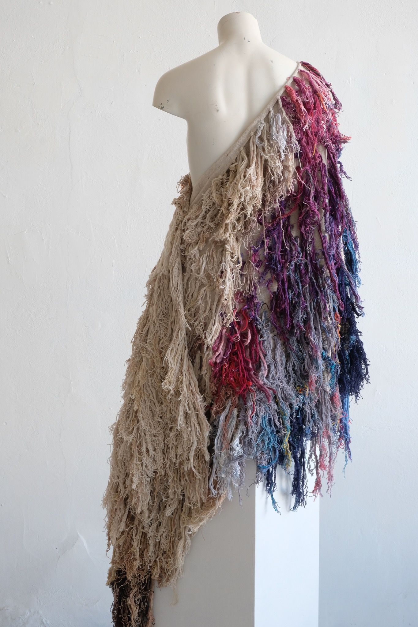 A rainbow, white and brown colored shawl, completely covered in fringe on a white mannequin, on a white pedestal, in a white room