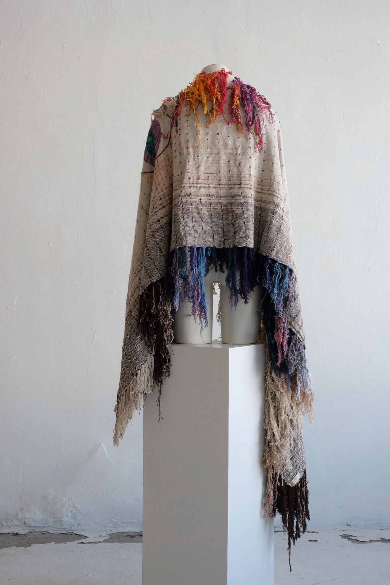 A rainbow, white and brown colored shawl, completely covered in fringe on a white mannequin, on a white pedestal, in a white room