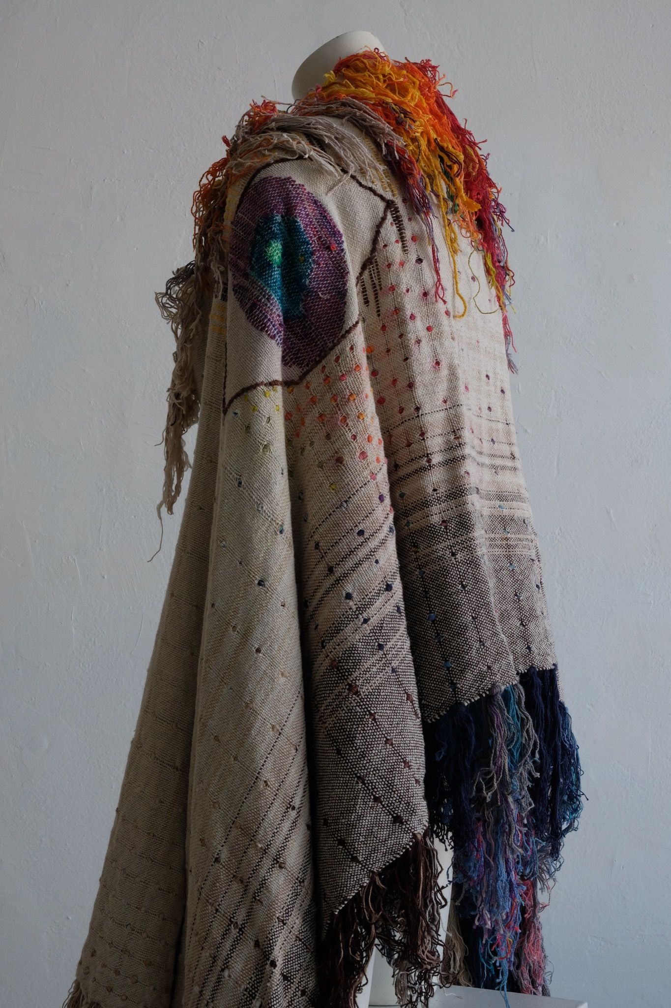 A rainbow, white and brown colored shawl, completely covered in fringe on a white mannequin, on a white pedestal, in a white room