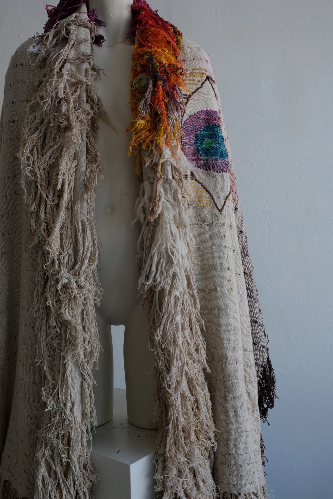 A rainbow, white and brown colored shawl, completely covered in fringe on a white mannequin, on a white pedestal, in a white room