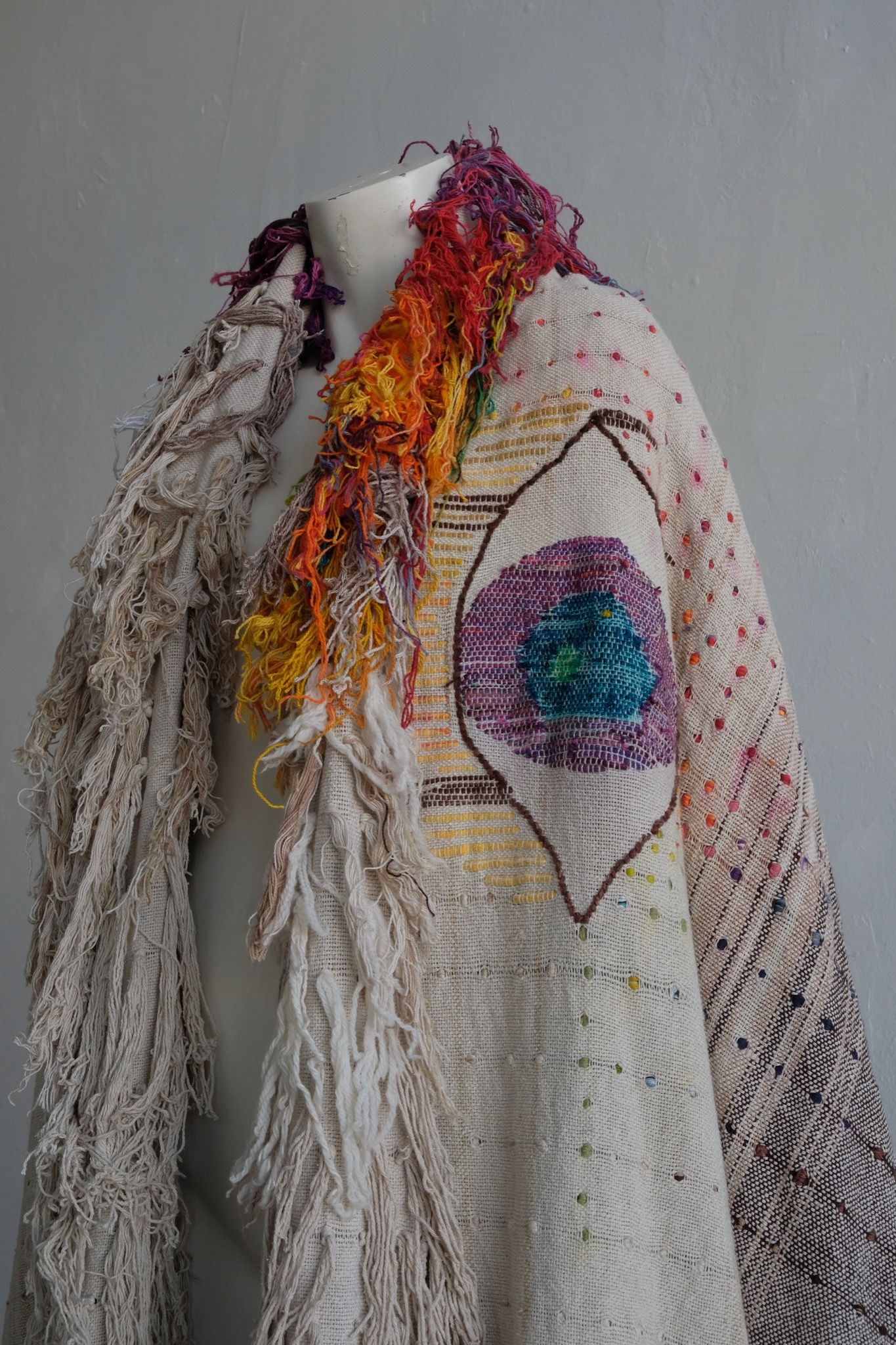 A rainbow, white and brown colored shawl, completely covered in fringe on a white mannequin, on a white pedestal, in a white room