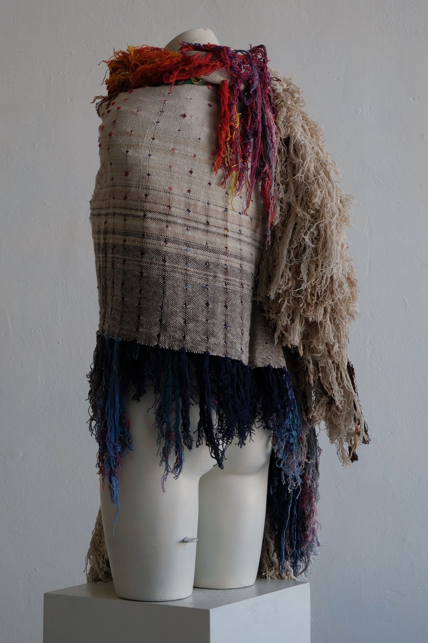 A rainbow, white and brown colored shawl, completely covered in fringe on a white mannequin, on a white pedestal, in a white room