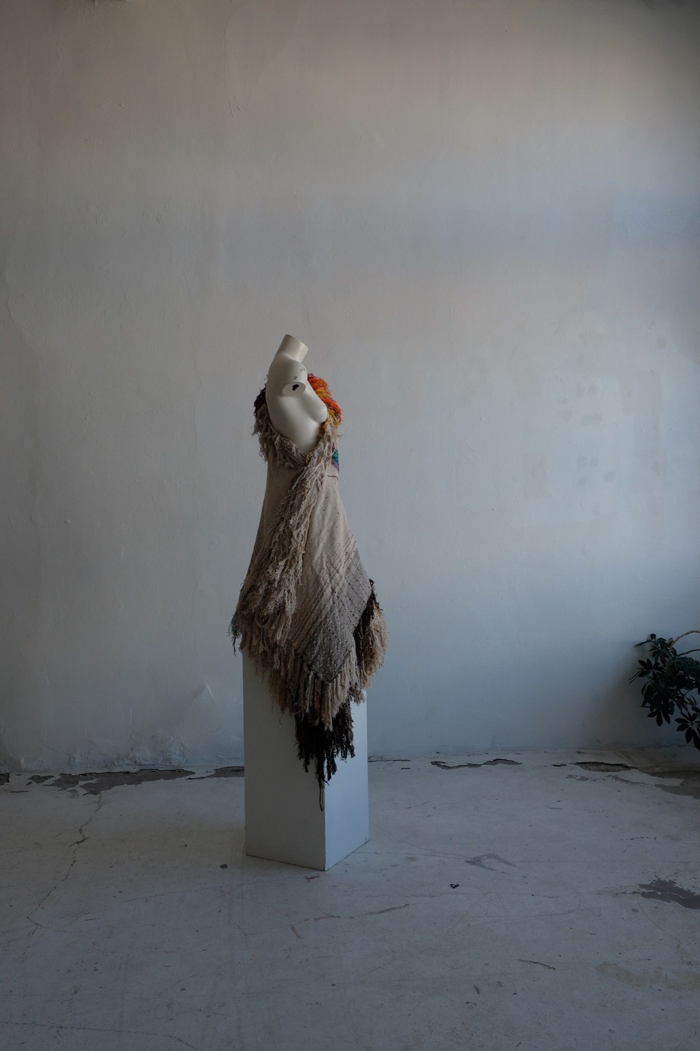 A rainbow, white and brown colored shawl, completely covered in fringe on a white mannequin, on a white pedestal, in a white room